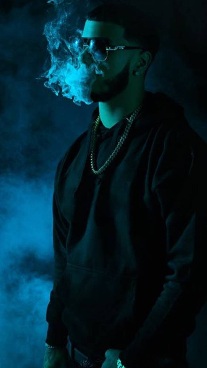 Anuel AA 黑暗風格造型 壁紙 — 一名身穿深色連帽衫、戴眼鏡的男子，在煙霧瀰漫的黑暗背景中呼出發光的藍色煙霧。