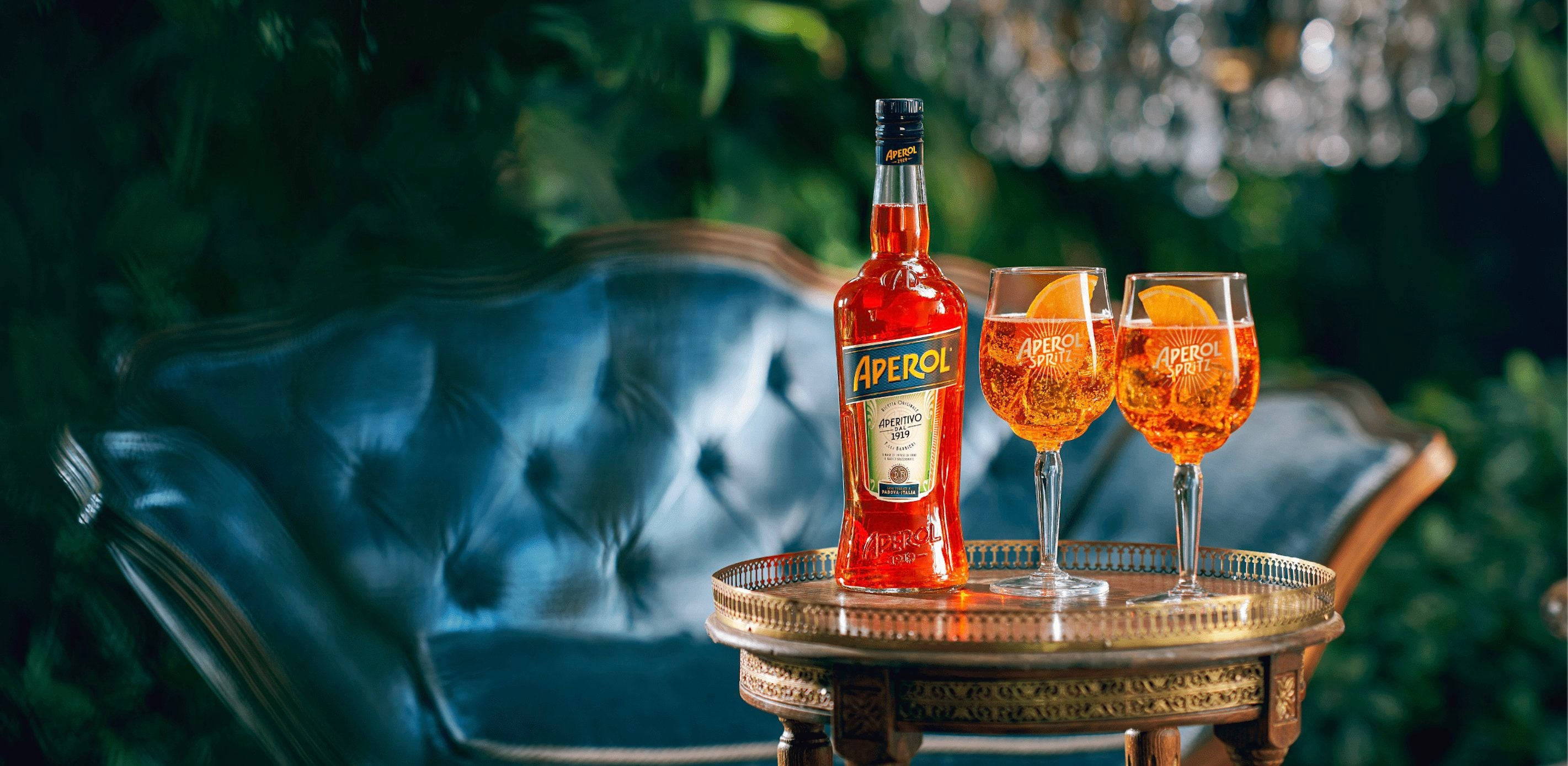 Aperol Spritz 飲料 壁紙 — 一瓶Aperol和兩杯帶有橙片的玻璃杯優雅地放在黃銅托盤上，前面是翠綠花園中的一張豪華藍色簇絨沙發。