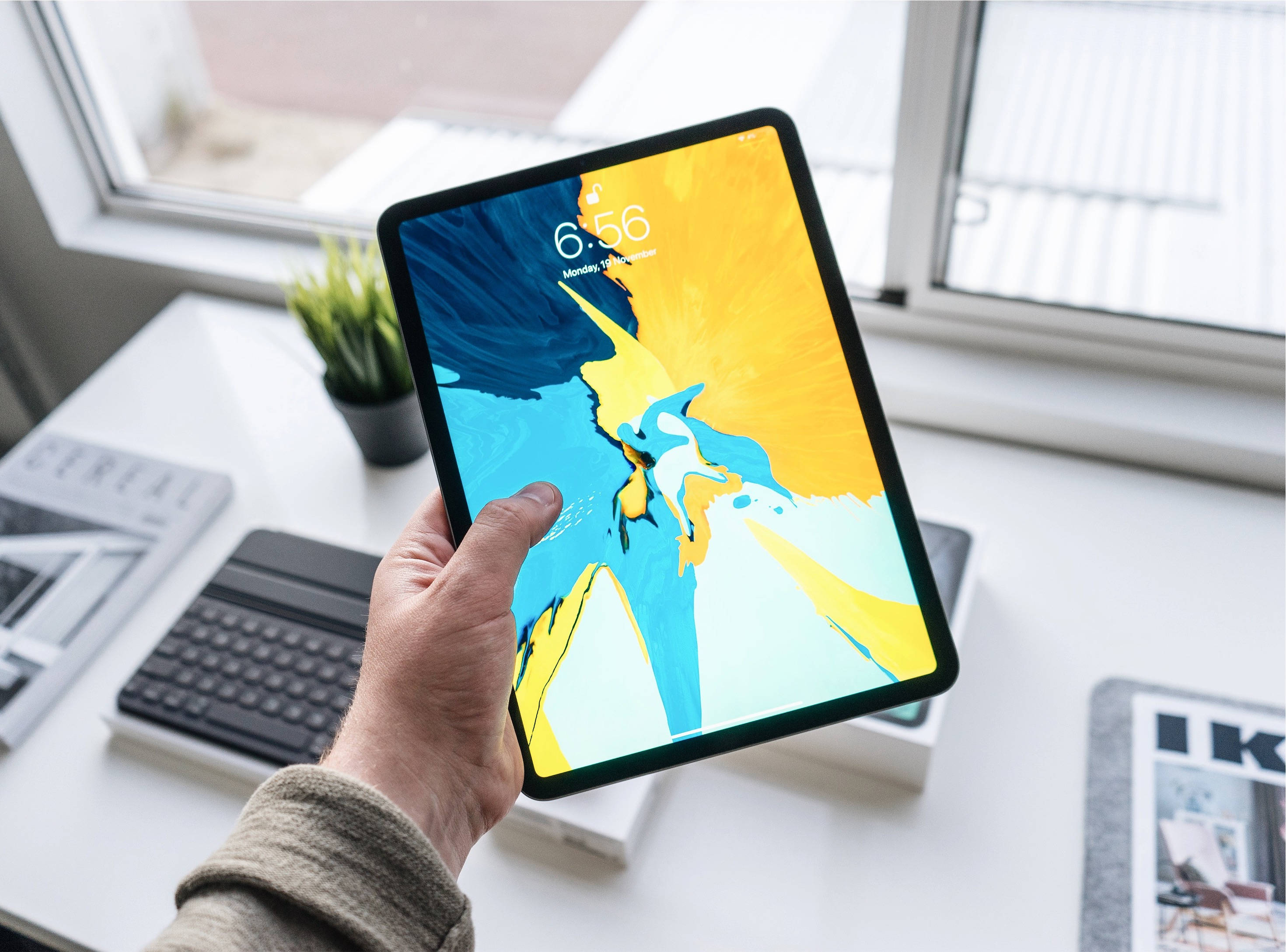 手持 Apple iPad Pro 的人物 壁紙 — 手拿著平板電腦，在明亮的現代辦公桌上顯示充滿活力的抽象壁紙，伴隨著窗戶光線。