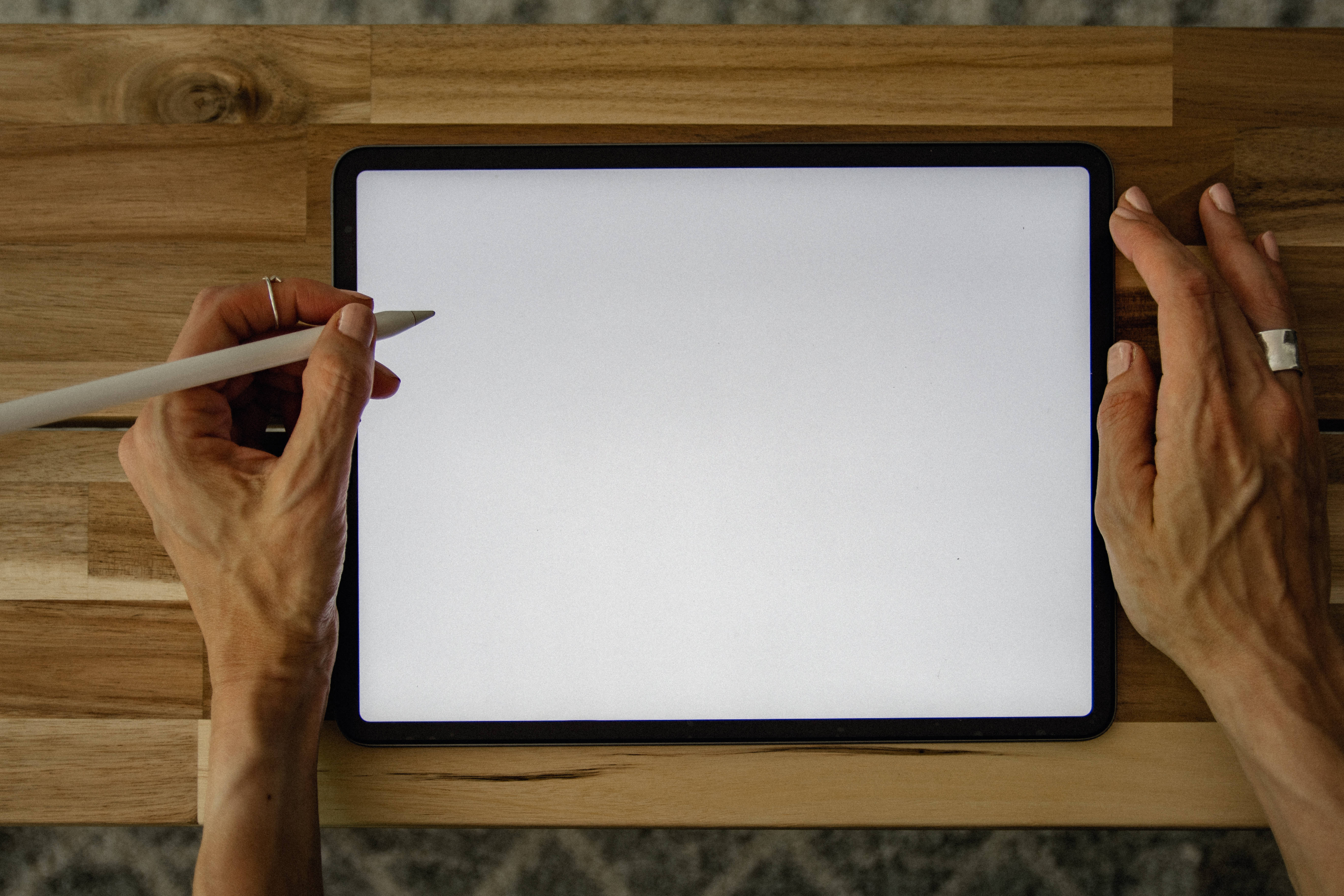 平板 Apple iPad Pro 壁紙 — 俯視角度，雙手在木桌上握著觸控筆，置於空白平板螢幕上方。