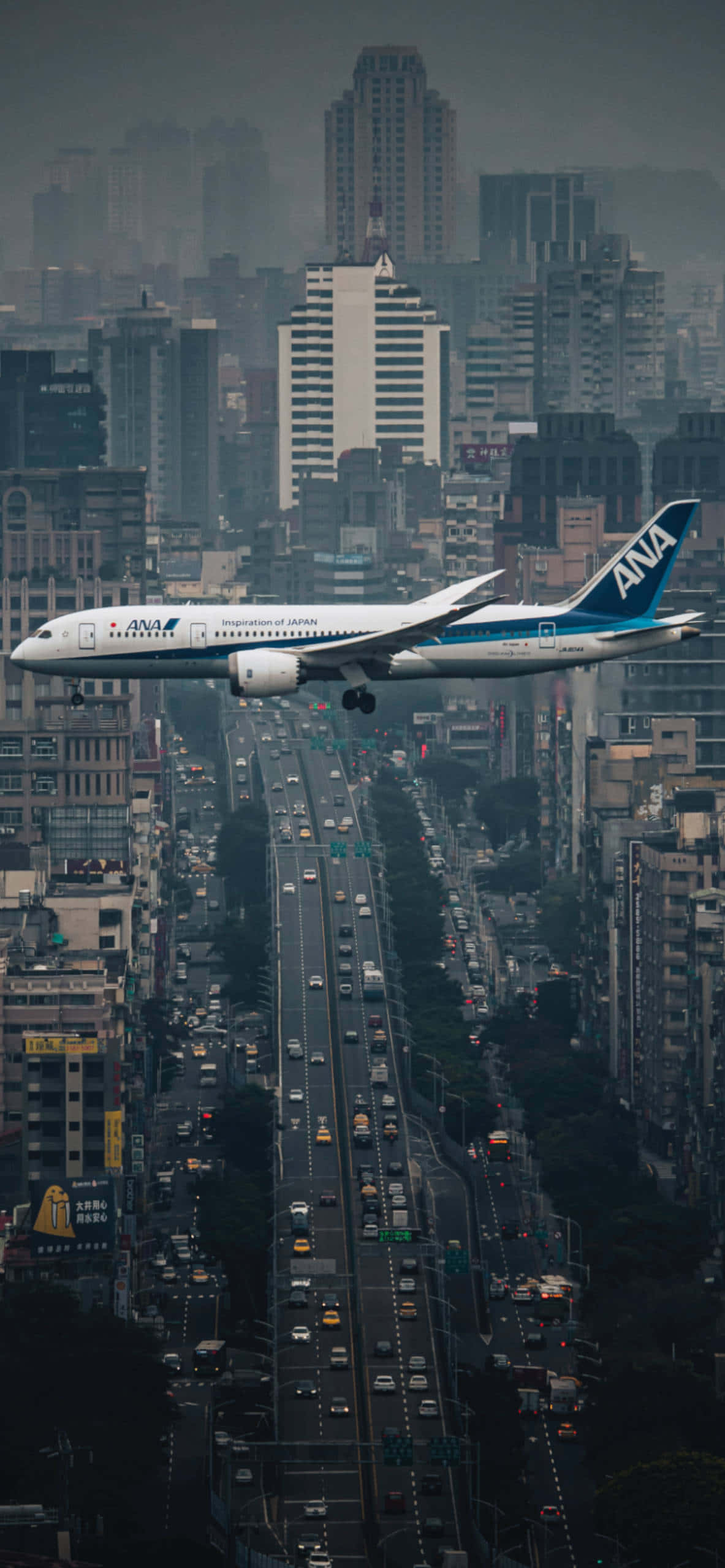 與最新Apple iPhone一同起飛 壁紙 — 一架全日空飛機在煙霞籠罩的天空下，低飛過一條車水馬龍、高樓林立的繁忙城市街道。