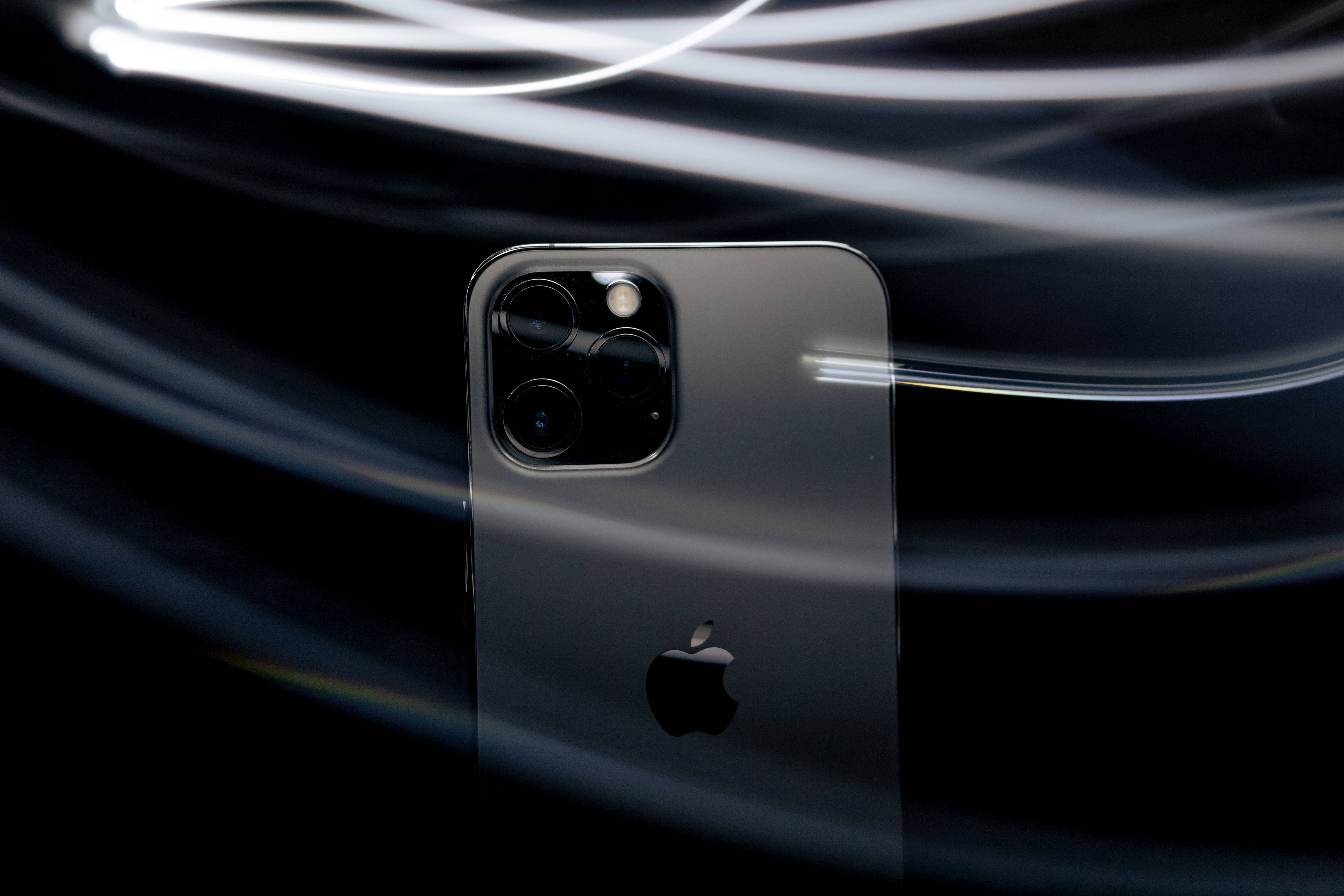 Apple Lidar 潮流 iPhone 壁紙 — 一部時尚的深灰色 iPhone 配備三鏡頭陣列，在黑色背景上與明亮的白色光軌動態漩渦形成對比。