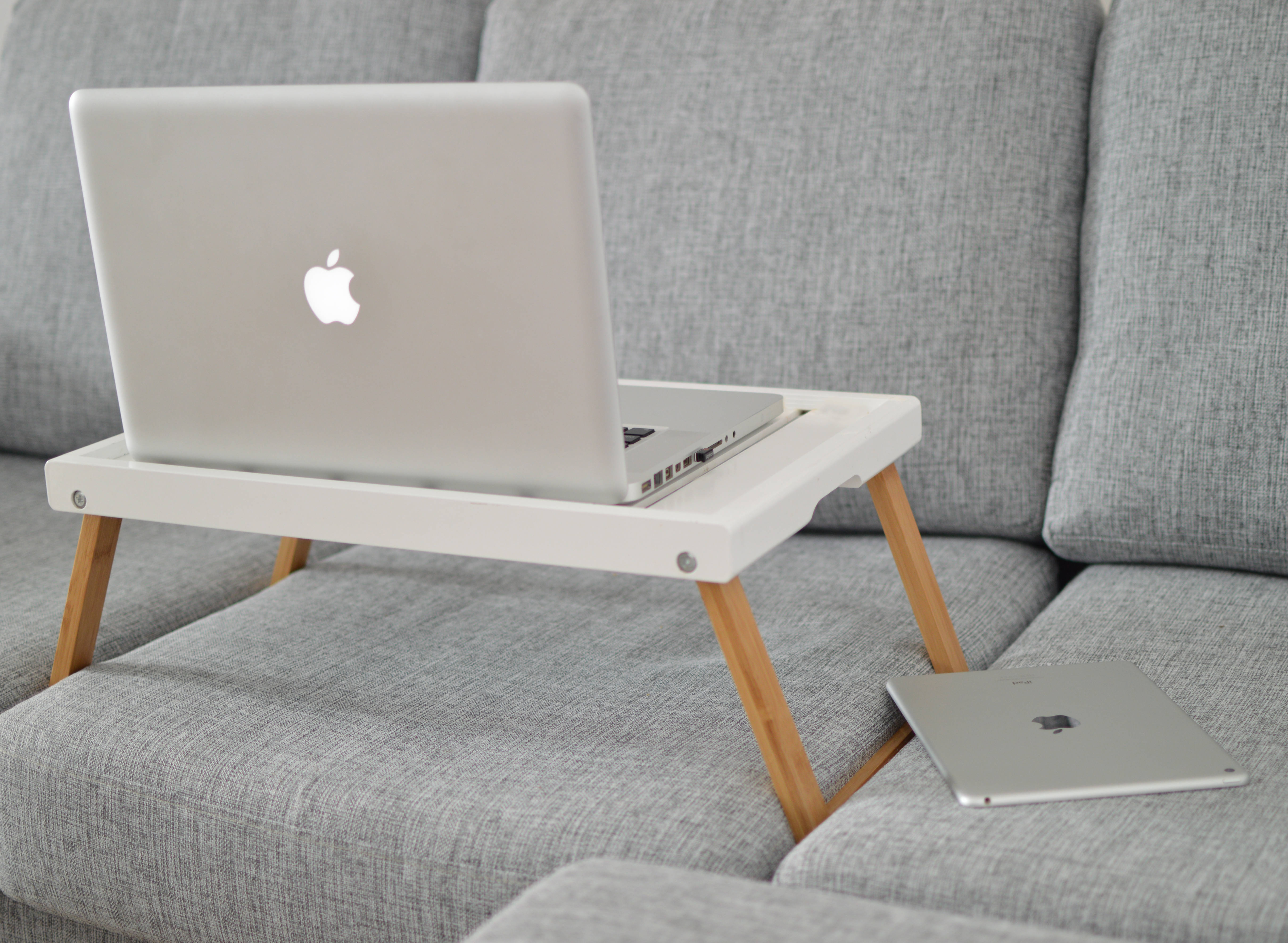 木質背景上的精緻 Apple MacBook Pro 壁紙 — 一部銀色手提電腦放在白色木製膝上桌上，桌子置於灰色布沙發上，旁邊有一部相配的平板電腦。