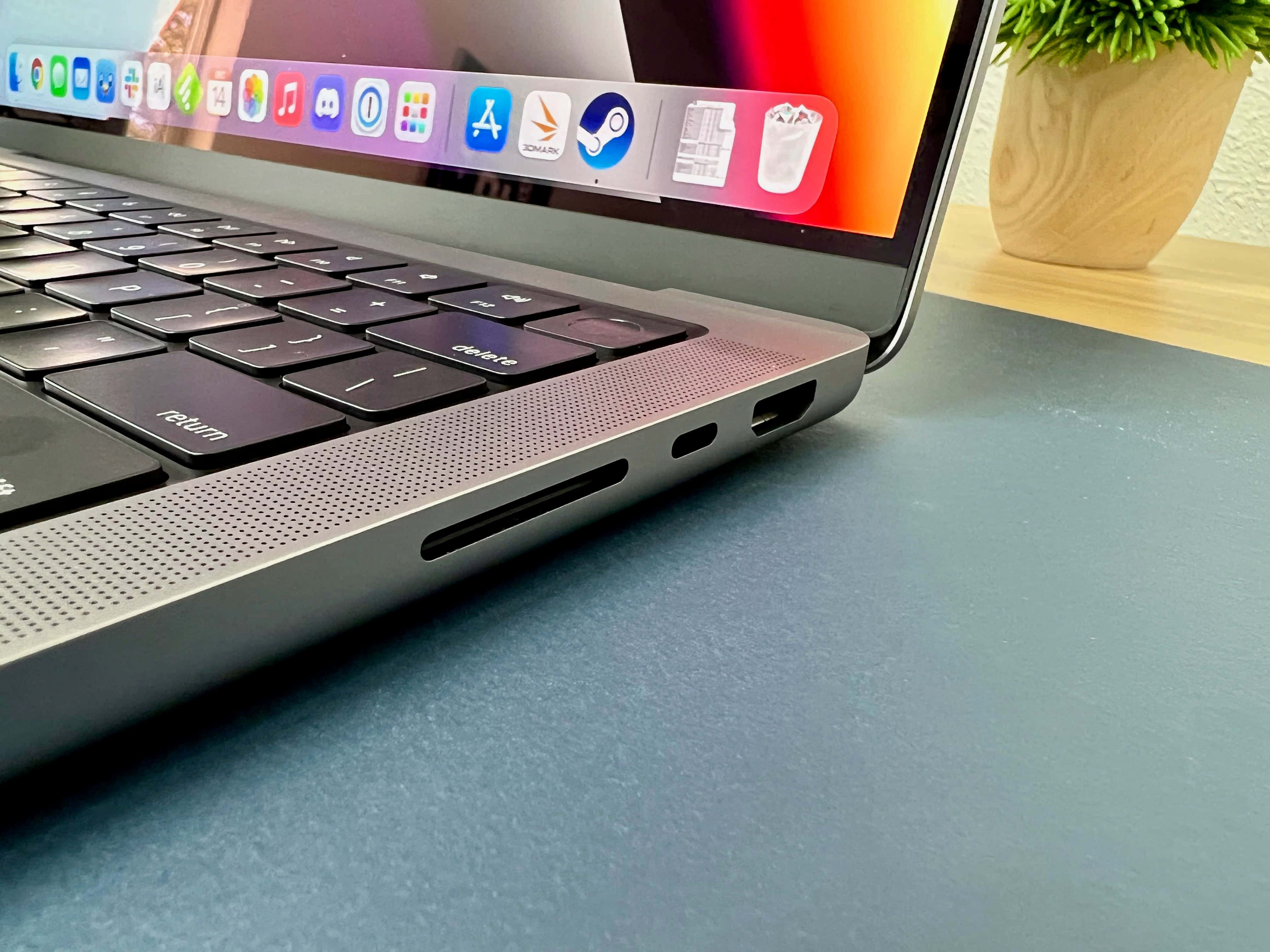 Apple MacBook，為強大工作體驗而設計 壁紙 — 藍色書桌上時尚銀色手提電腦的特寫，附近有盆栽植物，屏幕上顯示色彩繽紛的應用程式圖標。