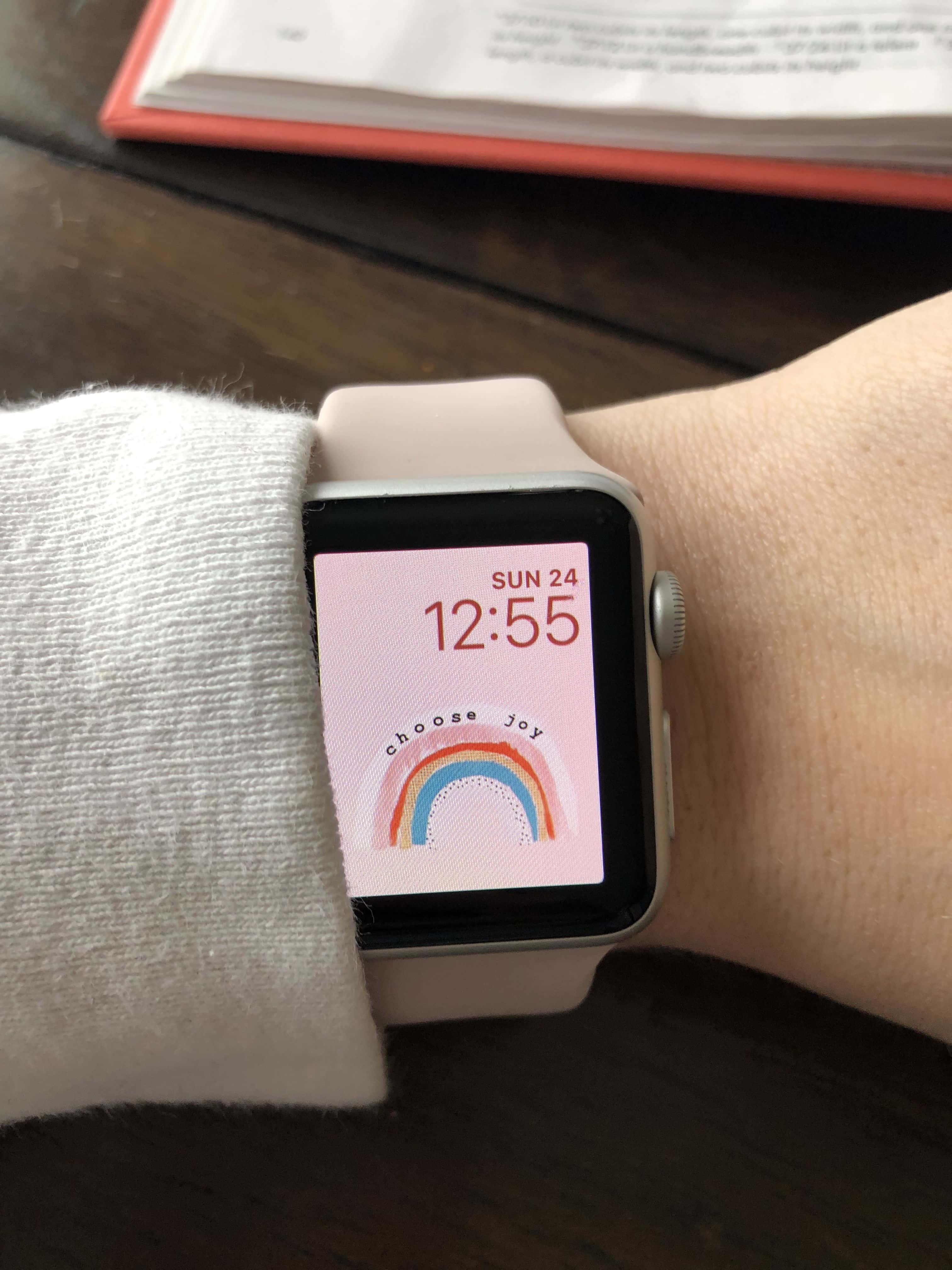 時尚實用的Apple Watch 壁紙 — 手腕佩戴智能手錶的特寫，粉紅色屏幕顯示彩虹和文字選擇快樂，背景為木桌。