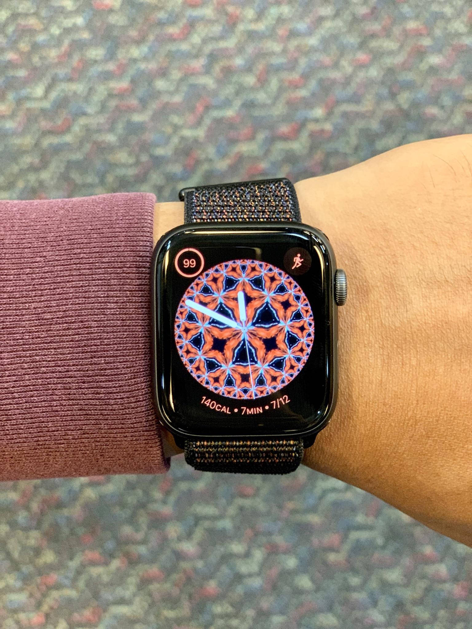 Apple Watch 彩虹旋轉錶盤 壁紙 — 手腕上智能手錶的特寫，在圖案地毯背景下顯示充滿活力的幾何鍛煉錶盤。