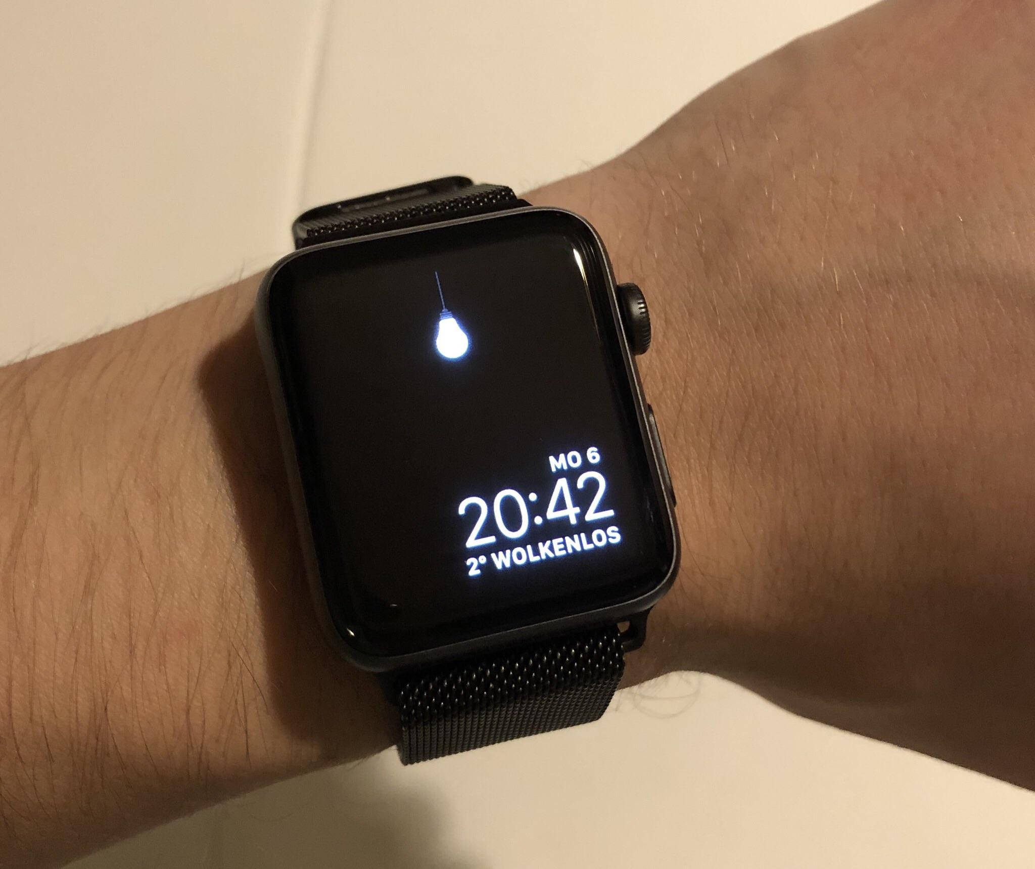 Apple Watch 懸掛燈泡介面 壁紙 — 手腕上黑色智能手錶的特寫，顯示時間、天氣和一個水滴圖標，背景為純色牆壁。