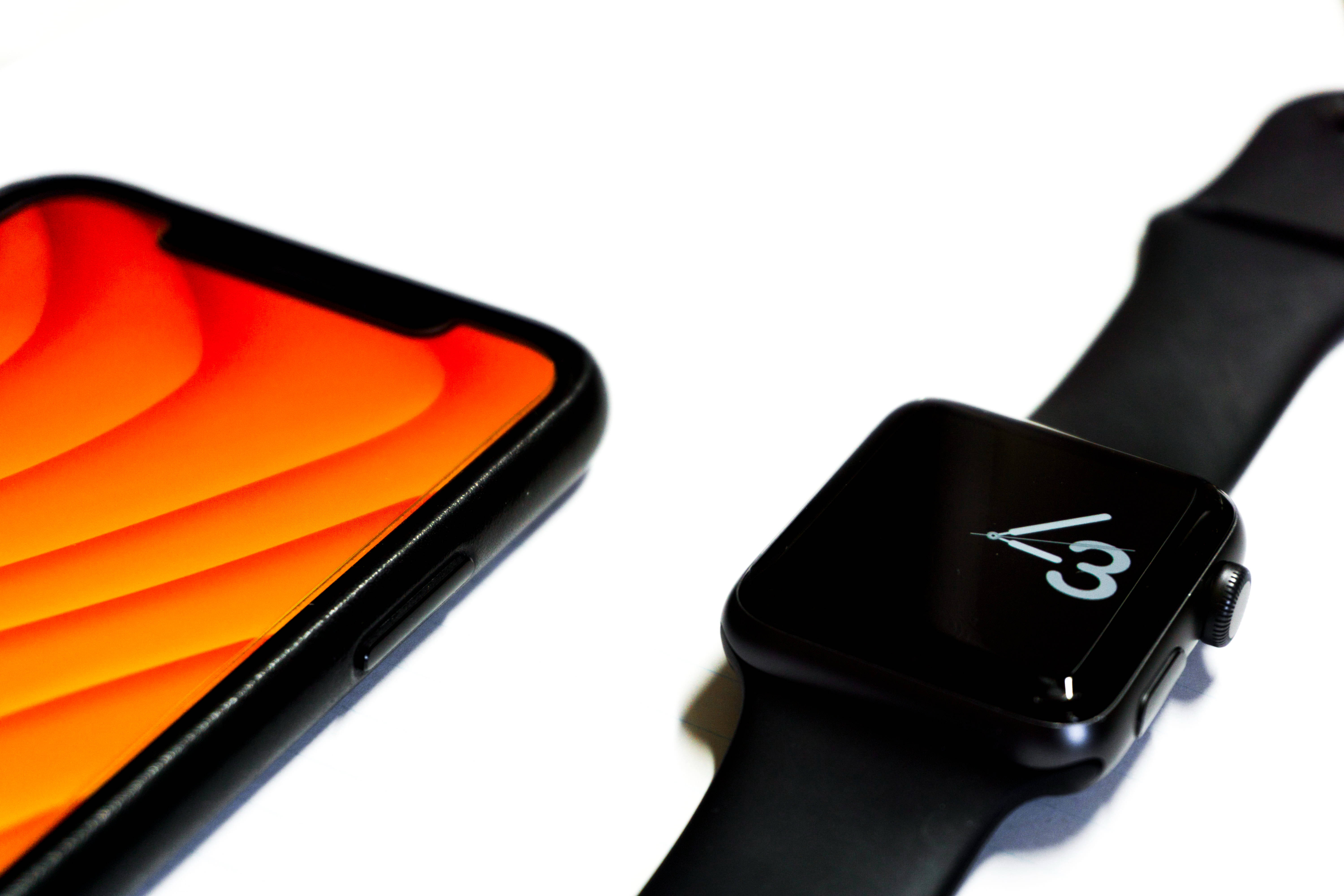 Apple Watch 最簡界面 壁紙 — 一部時尚的黑色智能手機帶有充滿活力的橙色漸變壁紙，旁邊放著一隻黑色Apple Watch，在簡約的白色表面上顯示時間4:30。