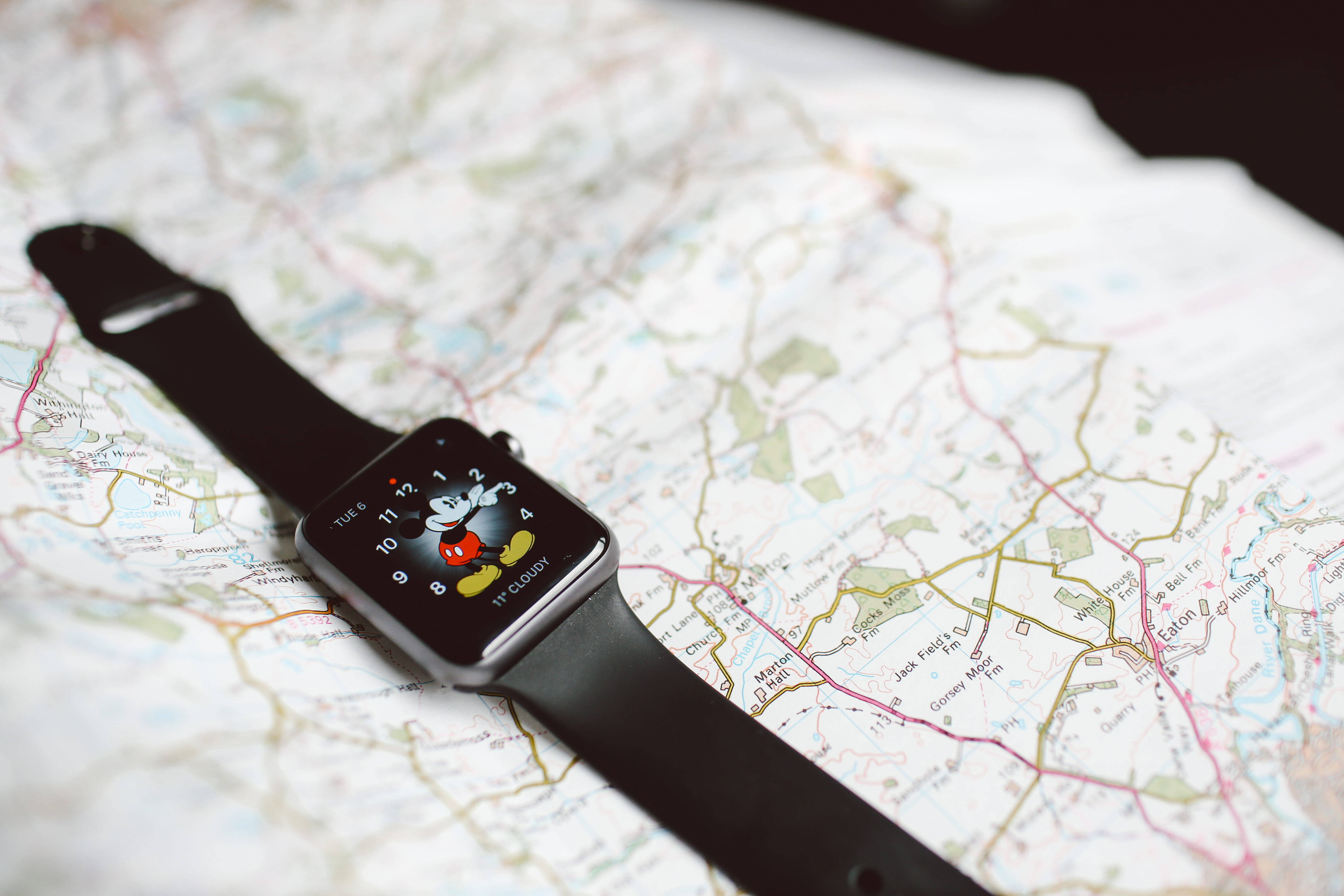 Apple Watch 米奇圖案界面 壁紙 — 一隻黑色智能手錶帶有米奇老鼠錶面，放在一張顯示道路和城鎮的詳細紙質地圖上。