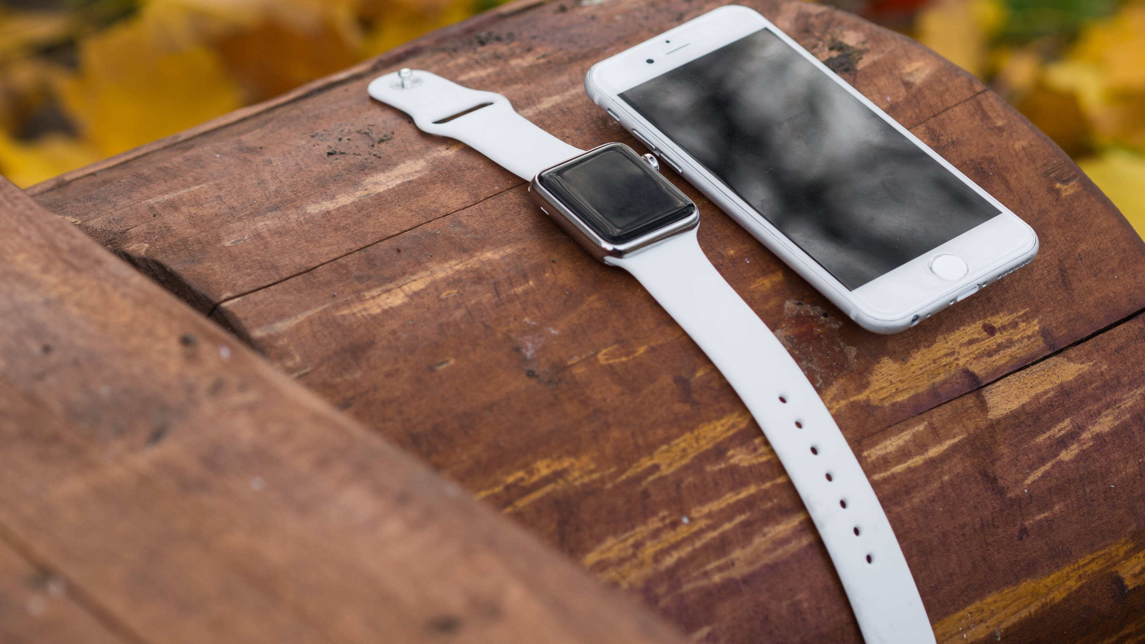 Apple Watch 經典白色款 壁紙 — 一隻白色智能手錶和智能手機放在經風化的木質表面上，背景有模糊的秋葉。