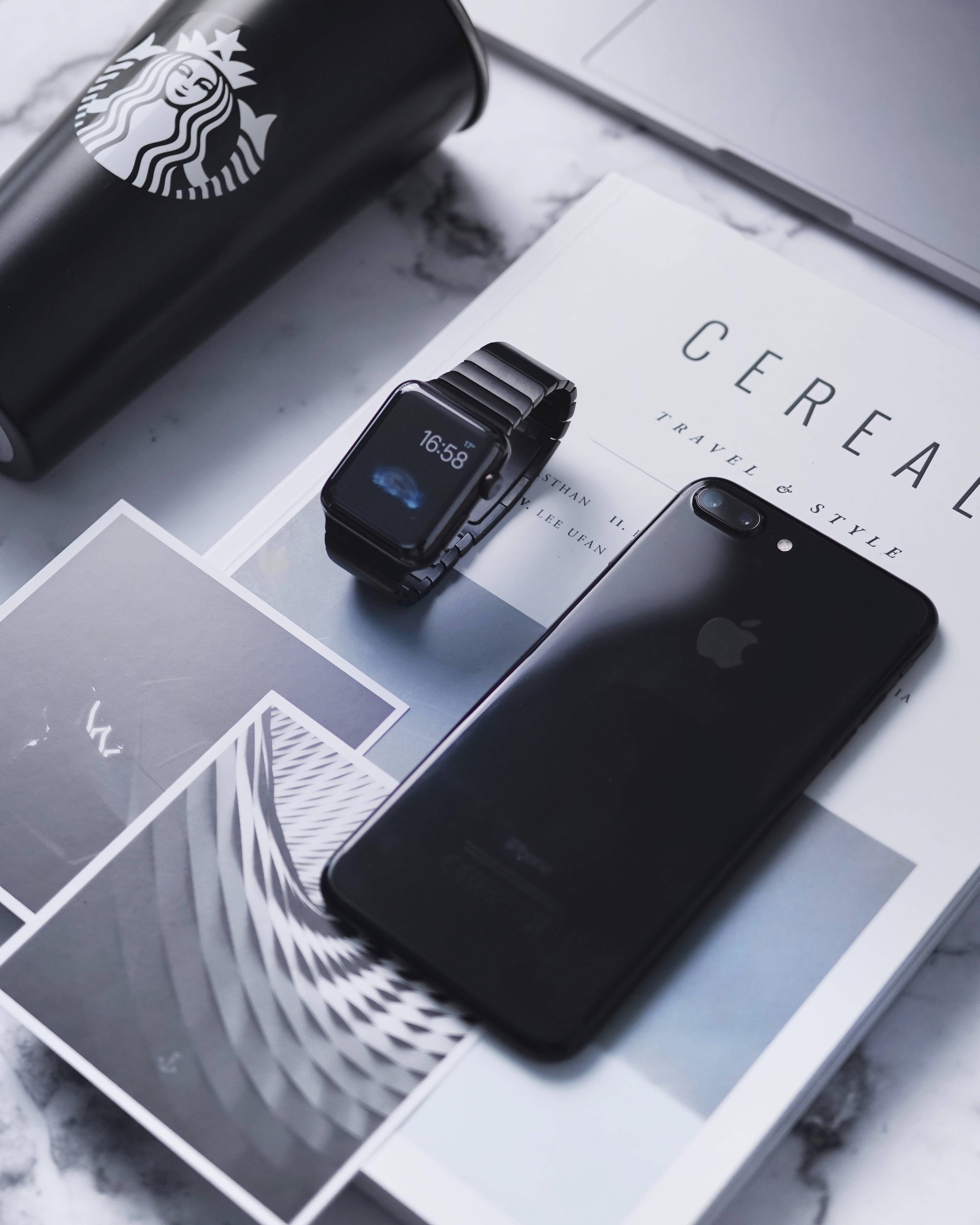 Apple Watch 與 Jet Black iPhone 壁紙 — 時尚黑色智能手錶和iPhone放在大理石表面的雜誌上，旁邊放著Starbucks杯。