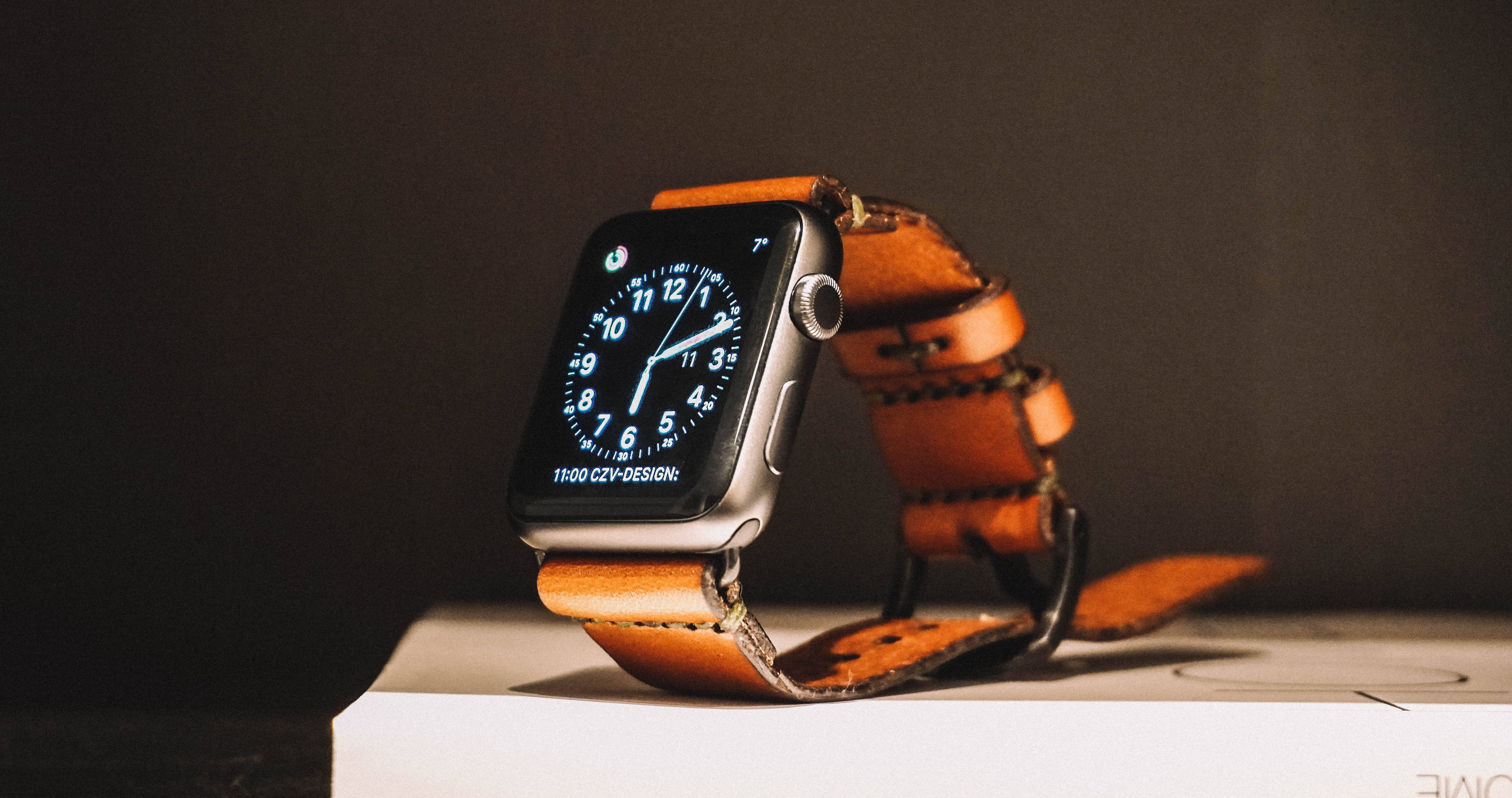 Apple Watch 配棕色皮革錶帶 壁紙 — 一款時尚的智能手錶配棕色皮革錶帶，放在白色盒子上，背景為深色。
