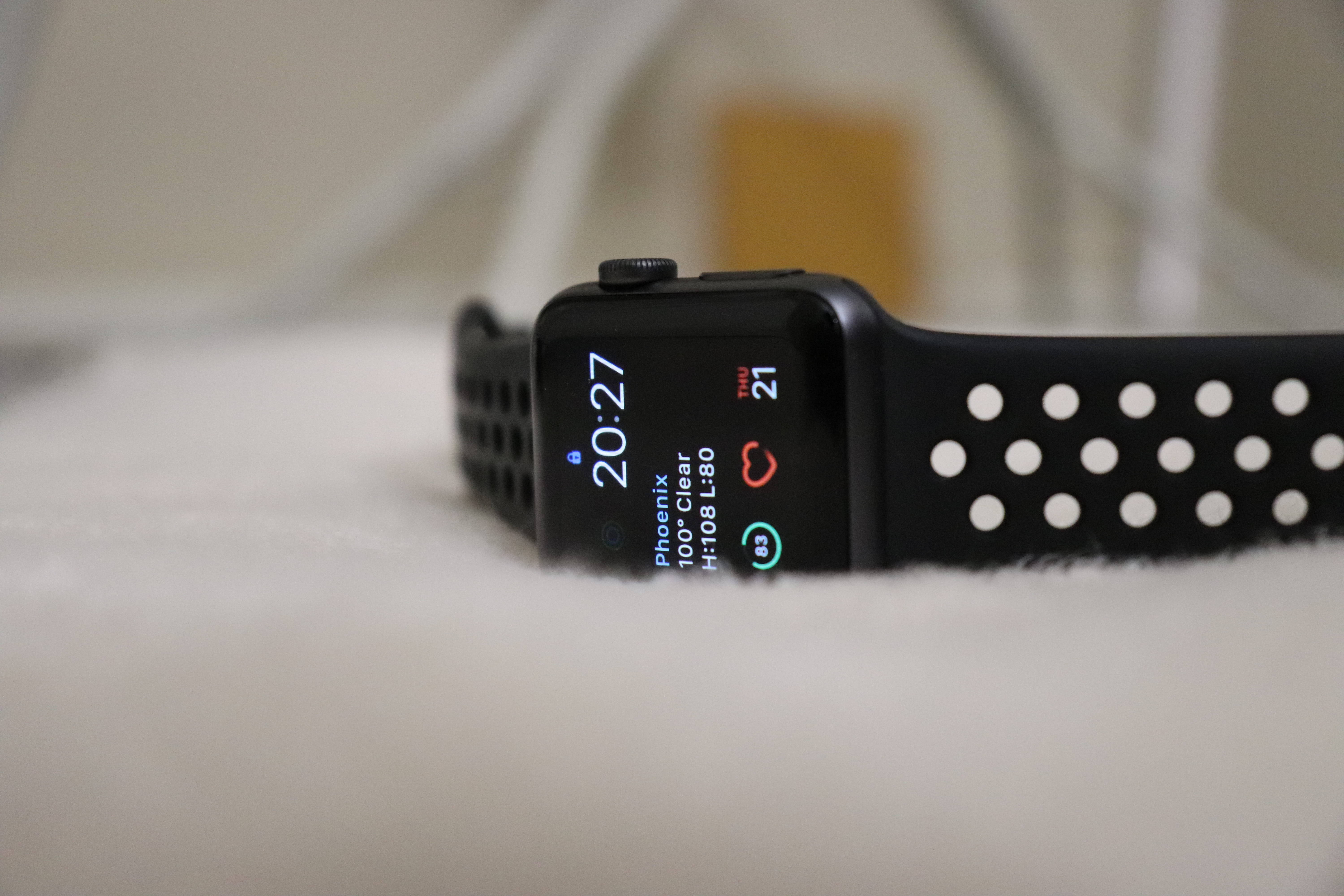 Apple Watch 配黑色錶帶 壁紙 — 黑色Apple Watch配穿孔錶帶，放置在柔軟的白色表面上，顯示時間、天氣和心率。