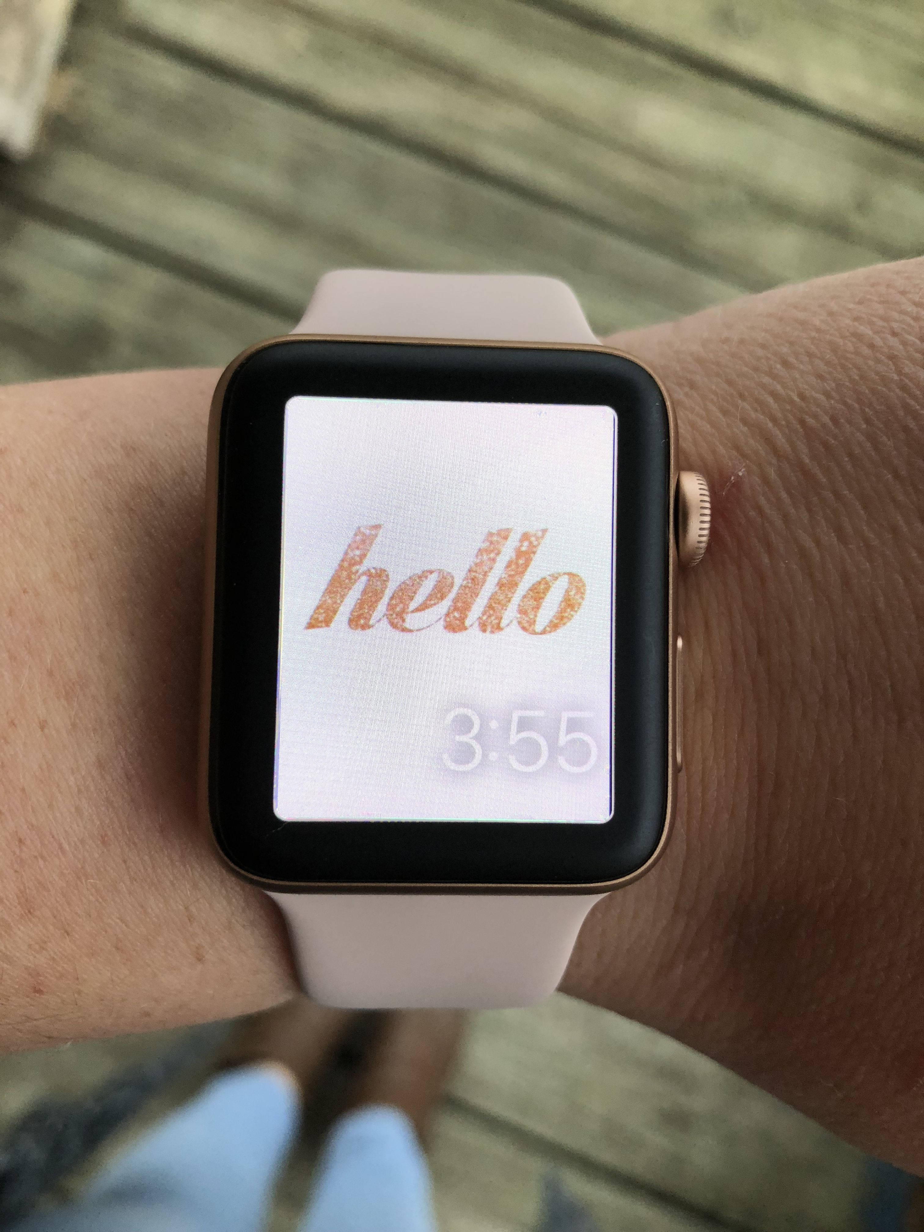 Apple Watch 霓虹閃粉 Hello 錶盤 壁紙 — 金色Apple Watch的特寫，配以淺粉紅色錶帶，在木質甲板背景上以玫瑰金色草書顯示單詞hello和時間3:55。