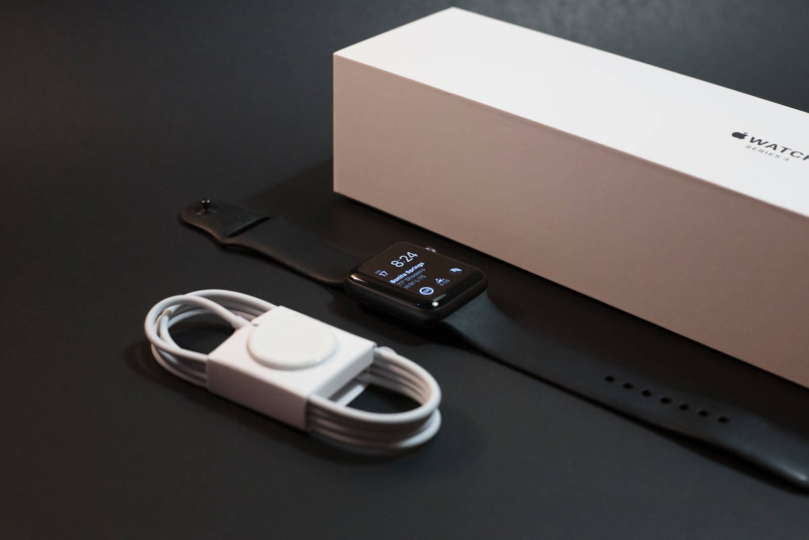 Apple智能手錶系列3 壁紙 — 一隻時尚的黑色Apple Watch Series 3連同白色充電線，放在其簡約白色盒子旁邊的深色表面上。