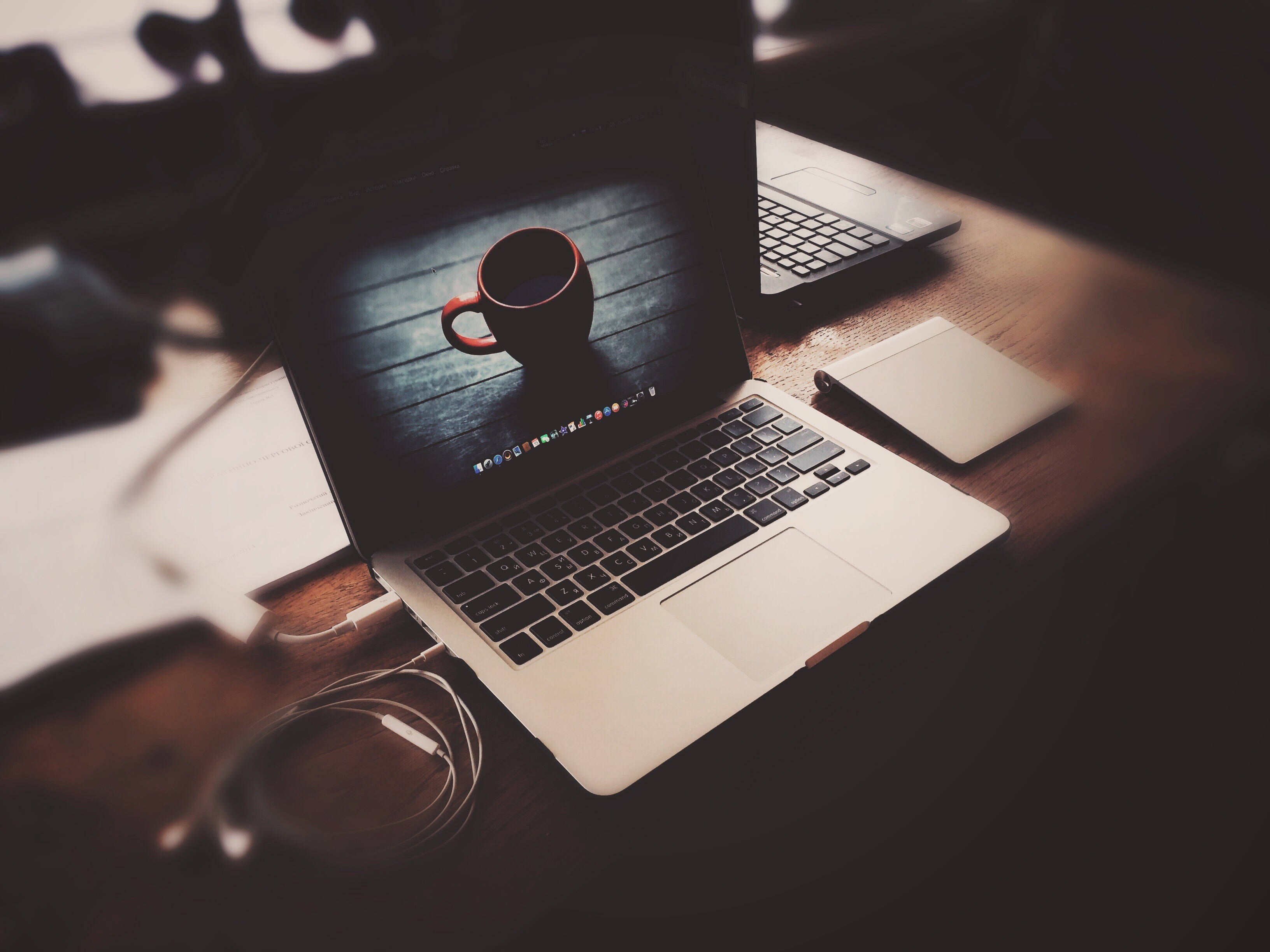 Apple產品：MacBook筆記本電腦與咖啡桌 壁紙 — 一部手提電腦放在木桌上，顯示咖啡杯壁紙，附近有電線和筆記本，在溫暖昏暗的燈光下。