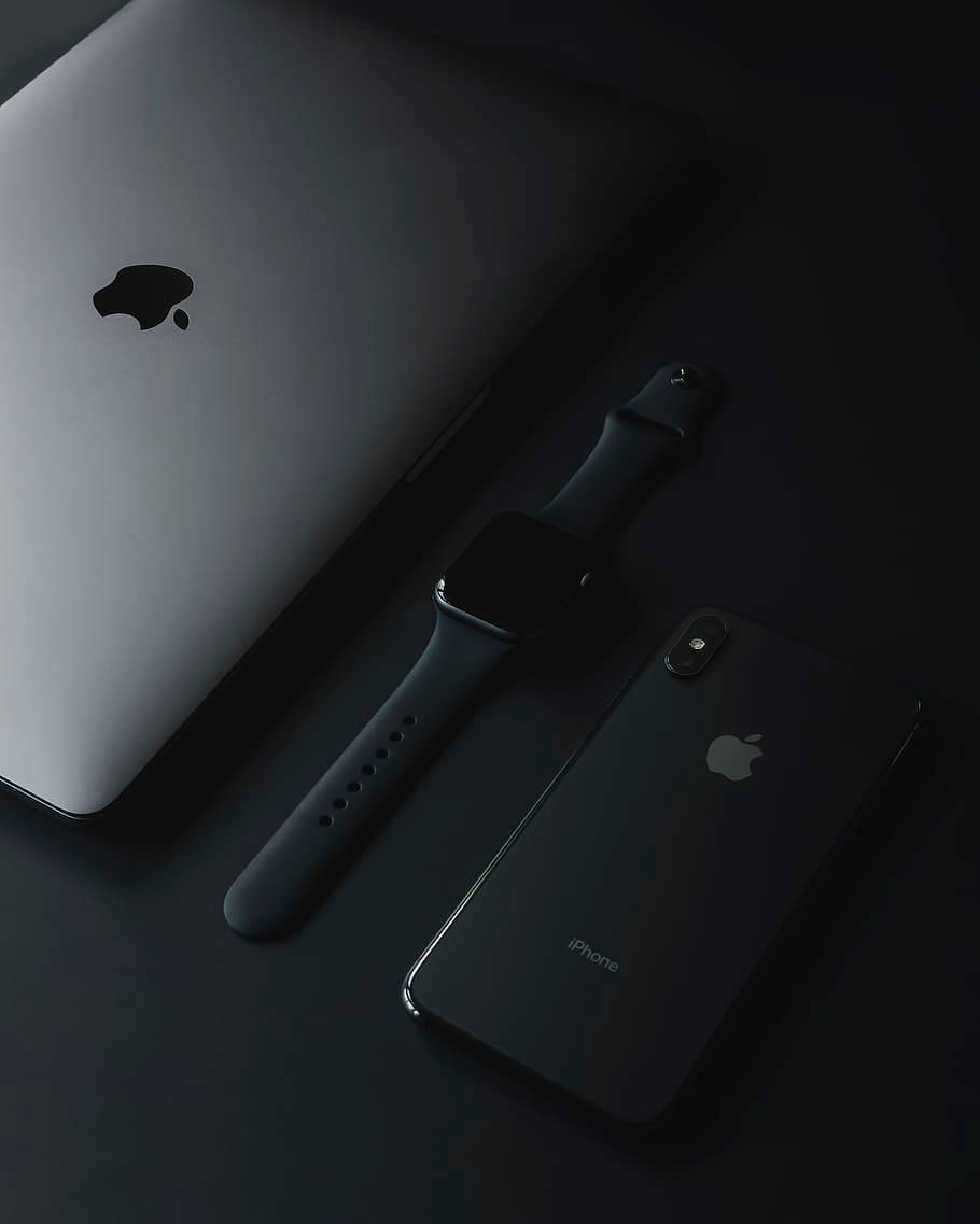 Apple產品：深色簡約風格 壁紙 — 一部時尚的銀色 MacBook 手提電腦、黑色 Apple Watch 和黑色 iPhone 在戲劇性燈光下排列在深色表面上。