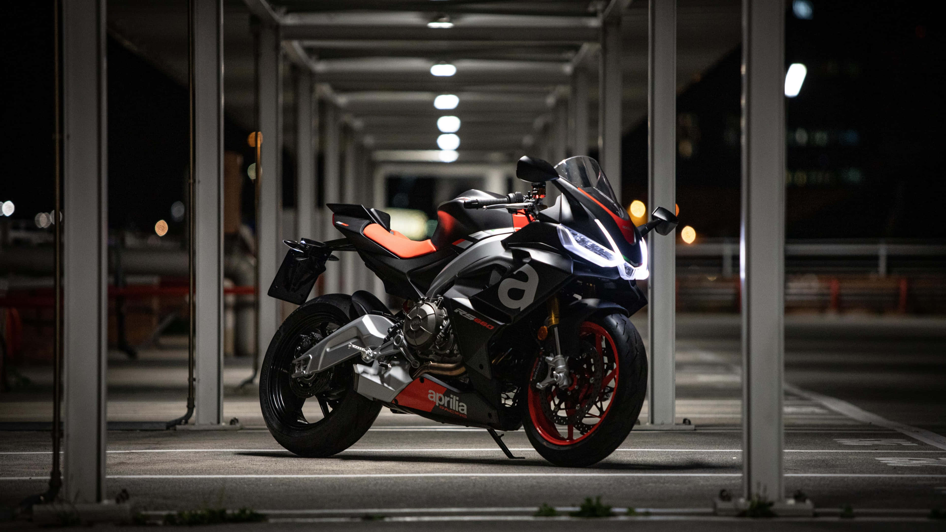 精美的Aprilia RSV4 RR機車背景 壁紙 — 一輛時尚的黑色和橙色Aprilia電單車在夜間有蓋的城市停車場中，在明亮的燈光下閃閃發光。