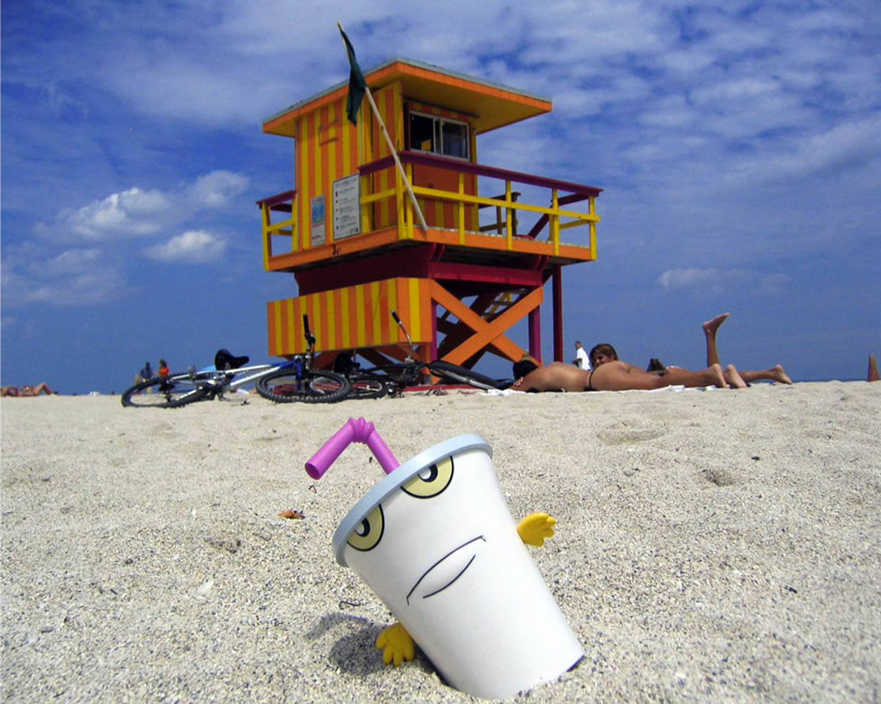 Aqua Teen Hunger Force 真實 Master Shake 壁紙 — 一個有眼睛和手臂的卡通杯從沙中冒出，前面是色彩繽紛的條紋救生塔，背景是陽光明媚的海灘。
