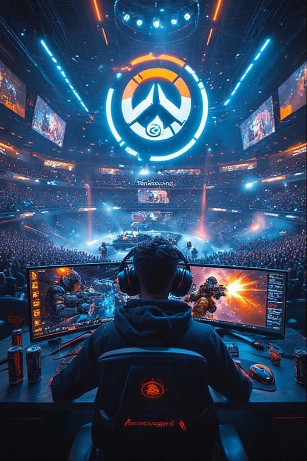 電競 arena 遊戲玩家壁紙 4K 壁紙 — 一位穿著連帽衫的玩家坐在桌前，面對兩個顯示《鬥陣特攻》遊戲畫面的顯示器，周圍是一個巨大的競技場，有發光的藍色和橙色燈光，上方有一個大型《鬥陣特攻》標誌。