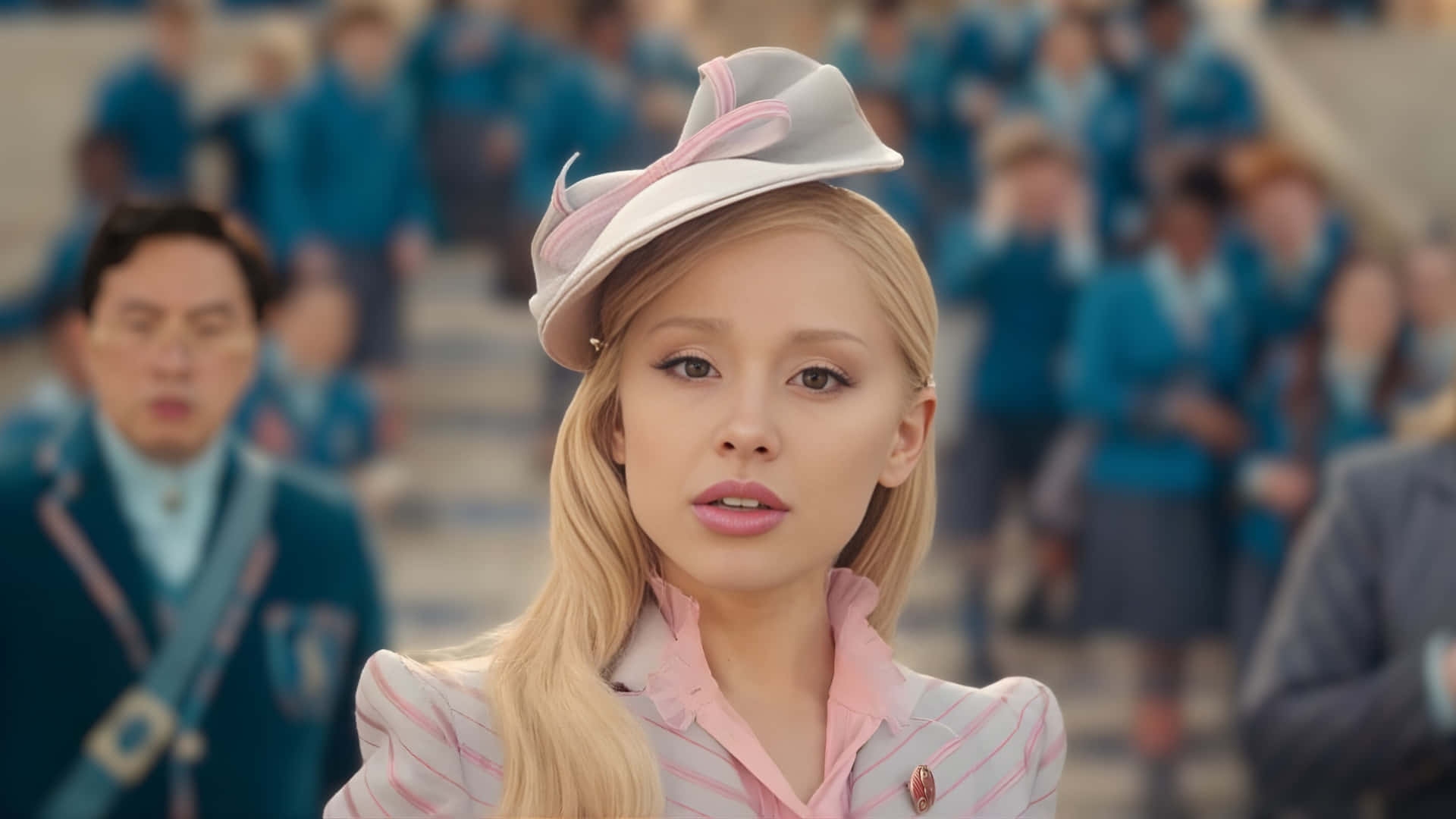  壁紙 — Ariana Grande 身穿粉白條紋制服，戴著相配的帽子，焦點清晰，而背景中模糊的同學則穿著藍色制服。