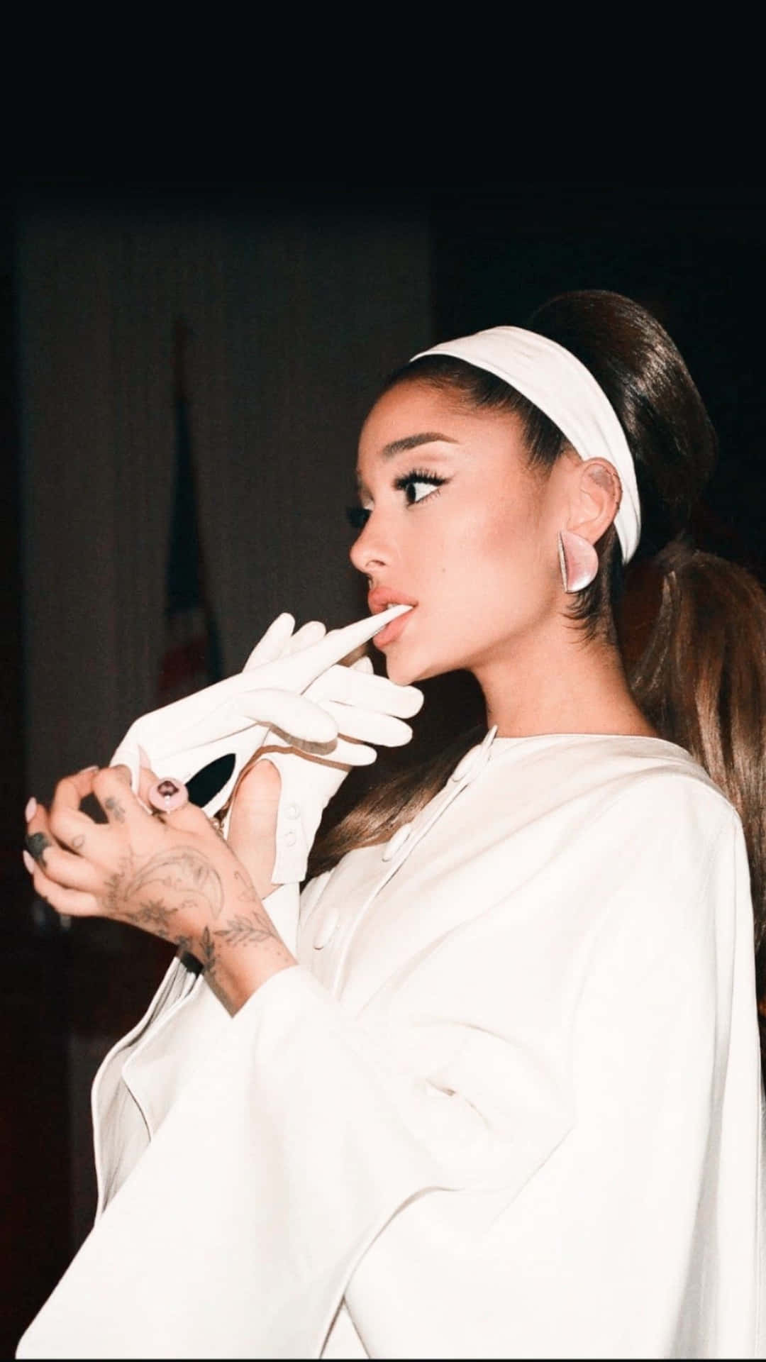  壁紙 — Ariana Grande 身穿時尚白色服裝和手套，在深色背景前將手指放在嘴唇上擺姿勢。