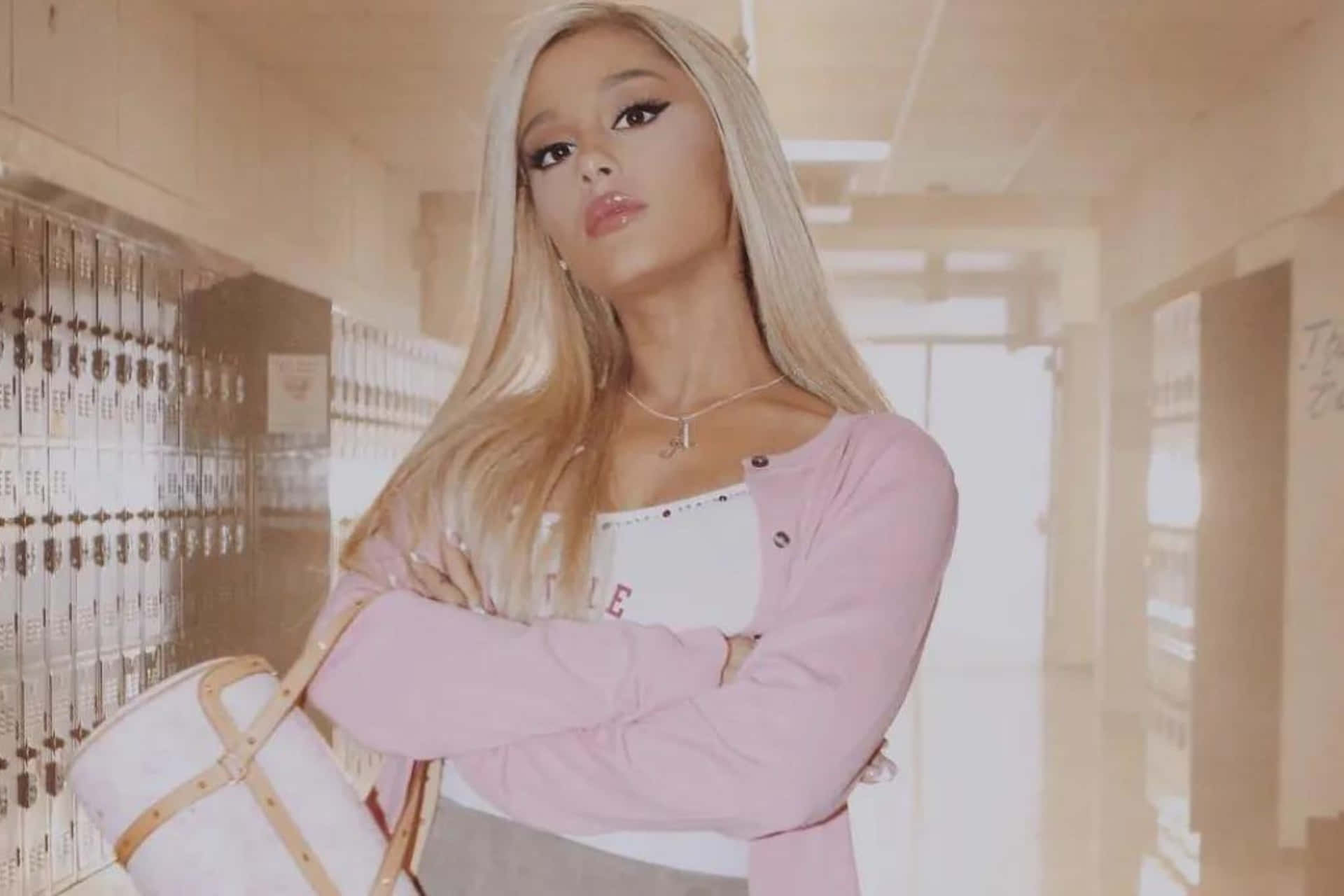  壁紙 — Ariana Grande 自信地站在陽光普照的學校走廊，雙臂交叉，身穿粉紅色開襟衫，手提白色手袋。