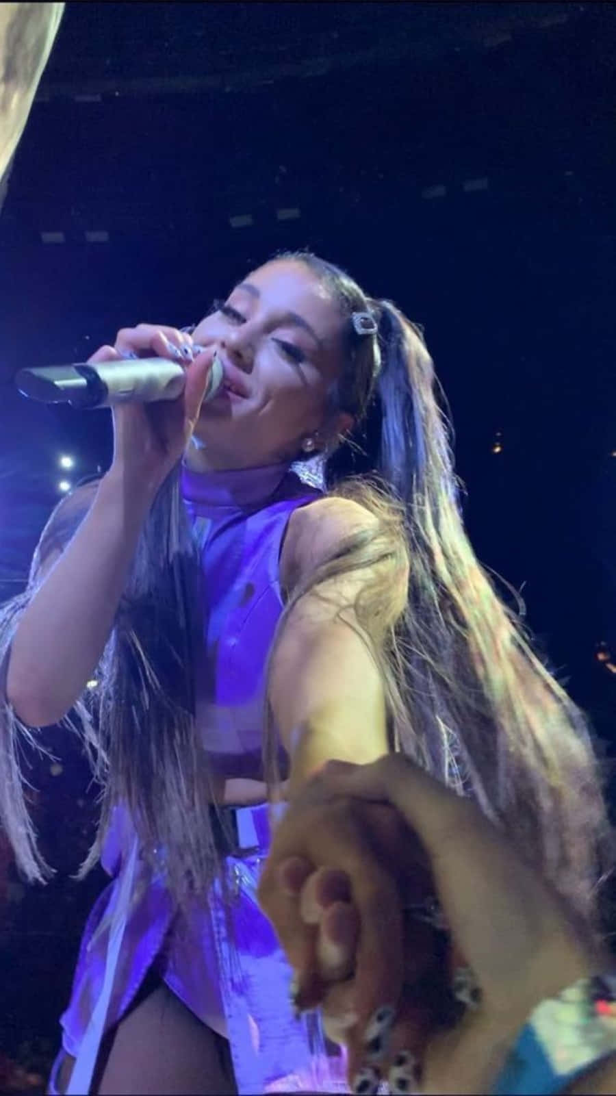 Aesthetic Ariana Grande 桌布 壁紙 — Ariana Grande 在紫色燈光下於舞台上對著咪高峰唱歌，同時在前景中握著一位粉絲的手。