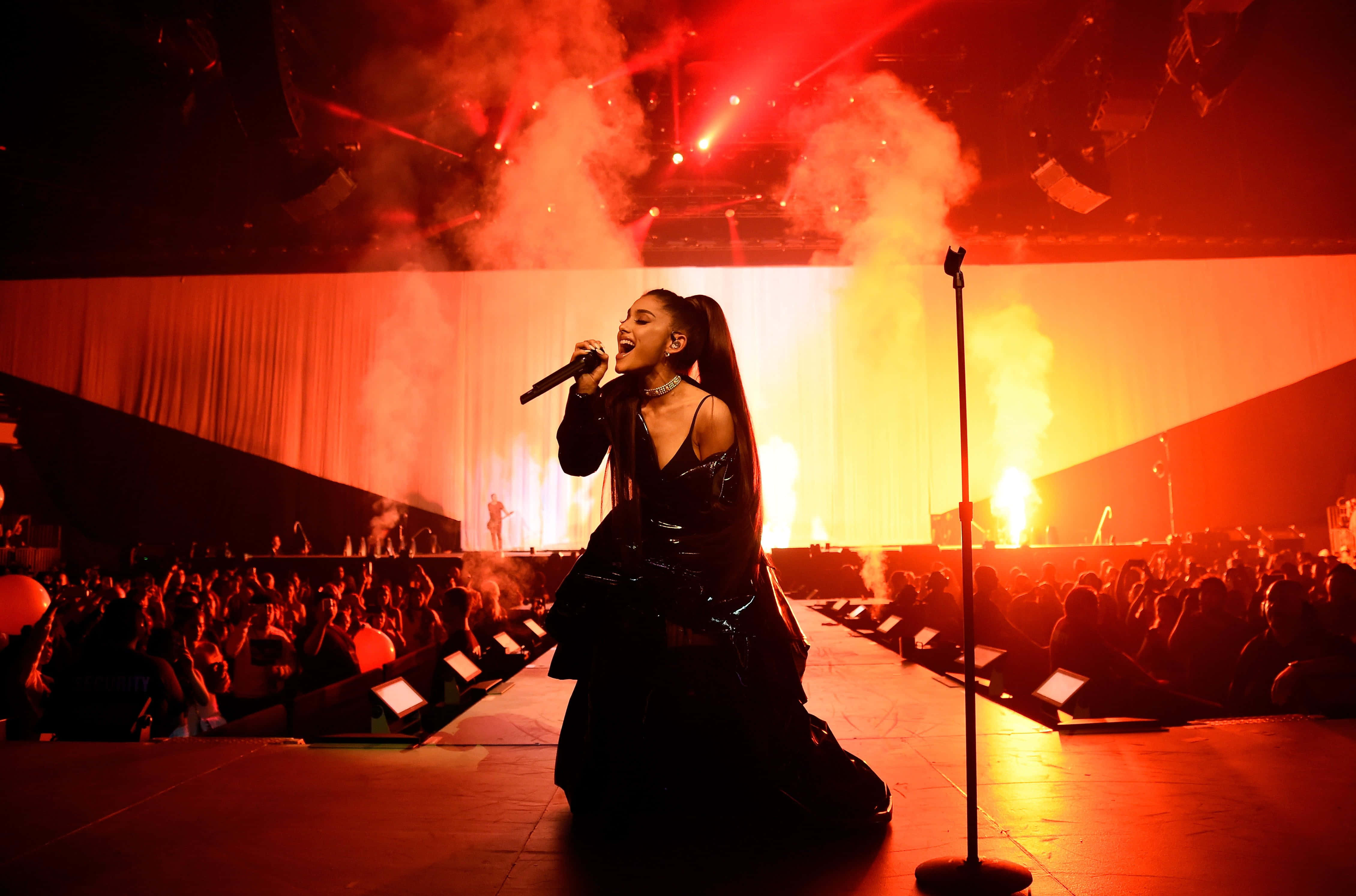Ariana Grande《Dangerous Woman》巡迴演唱會舞台背景 壁紙 — Ariana Grande 在煙霧和下方咆哮的人群中，於火紅燈光的舞台上熱情表演。