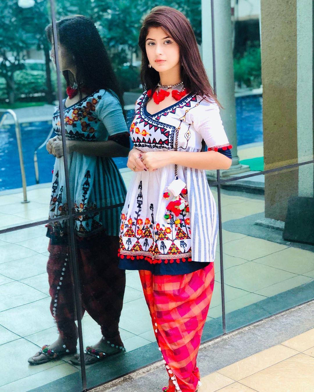 素雅的 Arishfa Khan 壁紙 — 一位年輕女性身穿白色刺繡庫爾塔和粉紅色格子褲，站在玻璃牆旁，玻璃牆反射出她和一個游泳池。