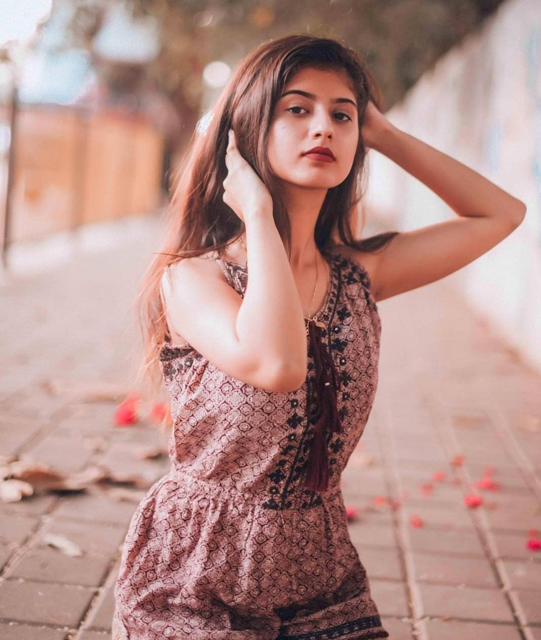 花卉風 Arishfa Khan 壁紙 — 一位長長棕色頭髮的年輕女子在溫暖的金色光線下，優雅地擺姿勢在散落著花瓣的瓷磚人行道上。