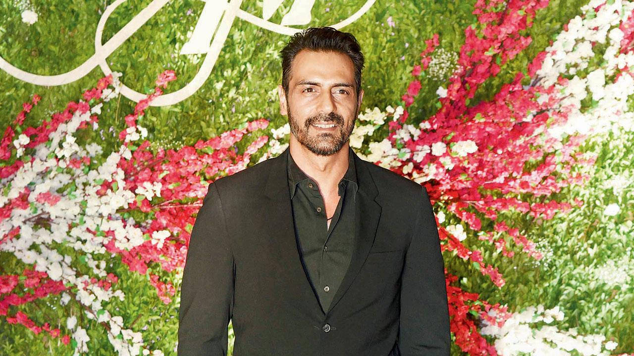Arjun Rampal 花紋背景造型 壁紙 — 一名身穿黑色西裝的男子自信地站在一面充滿粉紅色、白色和綠色花朵的茂盛花牆前。