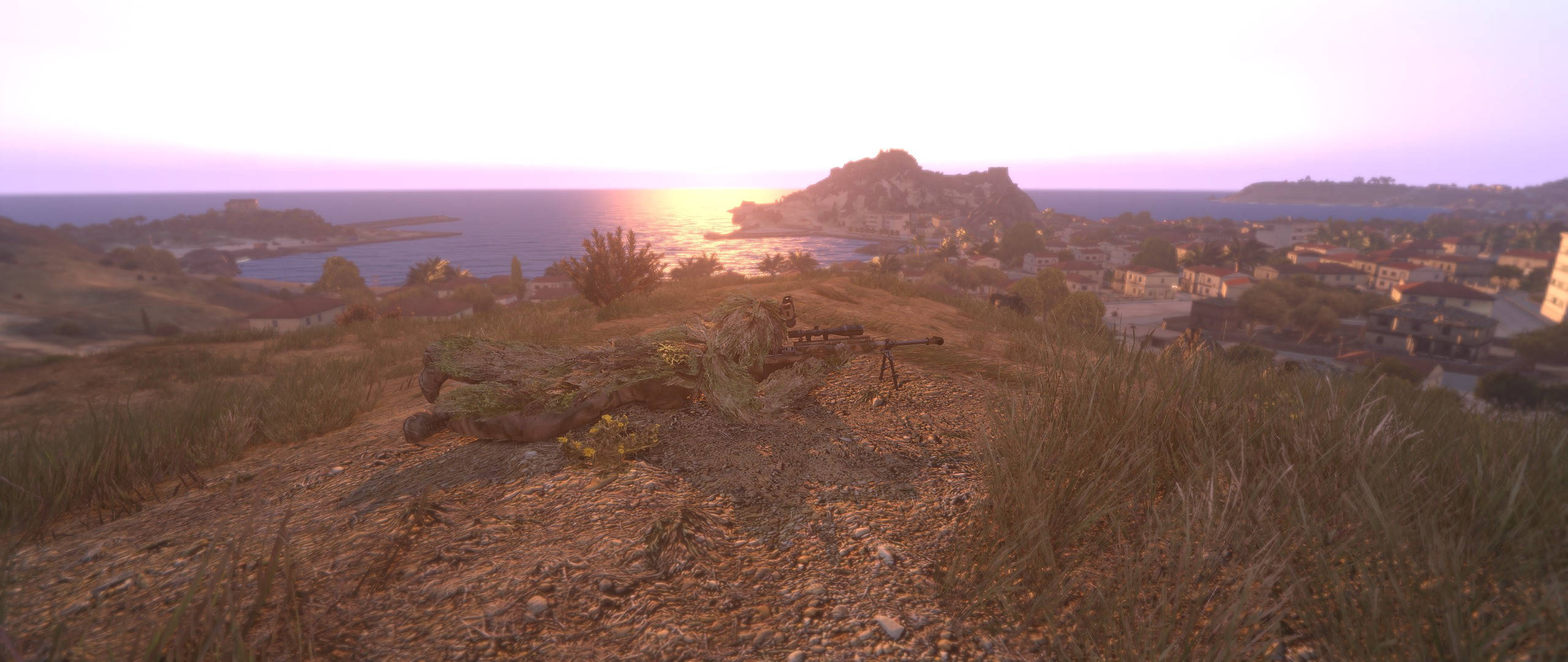 Arma 3 士兵穿越崎嶇地形 壁紙 — 一名身穿迷彩服的士兵俯臥在長滿草的山丘上，俯瞰著日落時分的沿海小鎮，溫暖的金色光芒灑落。