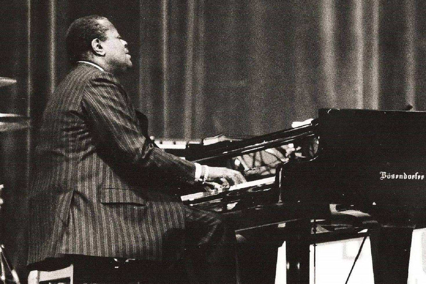 Art Tatum 在 Cafe Society 夜總會演出 壁紙 — 一位身穿細條紋西裝的爵士鋼琴家在燈光昏暗的舞台上，身後有窗簾，熱情地彈奏著三角鋼琴。