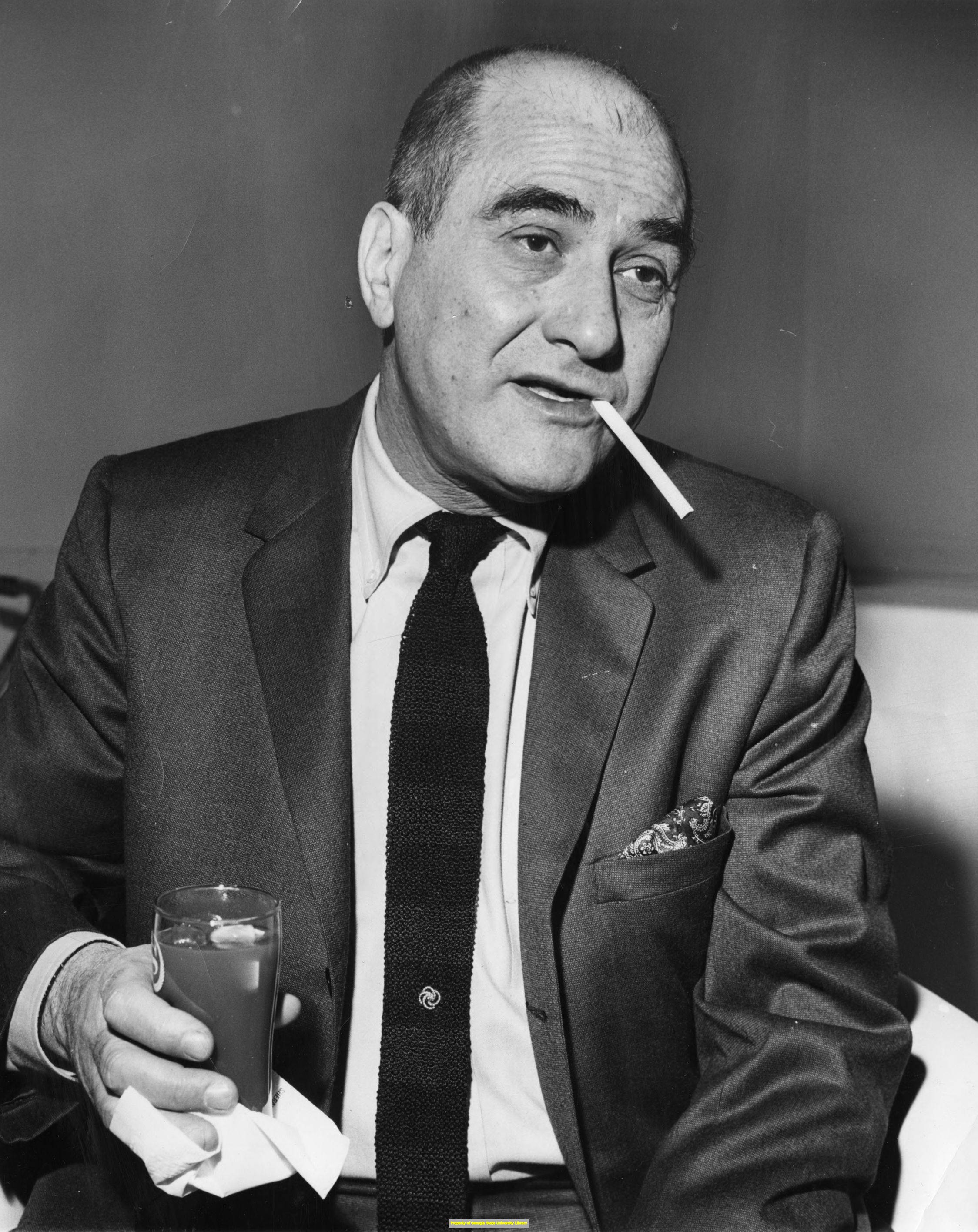 傳奇爵士樂手 Artie Shaw 沉思抽菸 壁紙 — 一個穿著西裝的男人嘴裡叼著煙，手持飲品的黑白肖像。