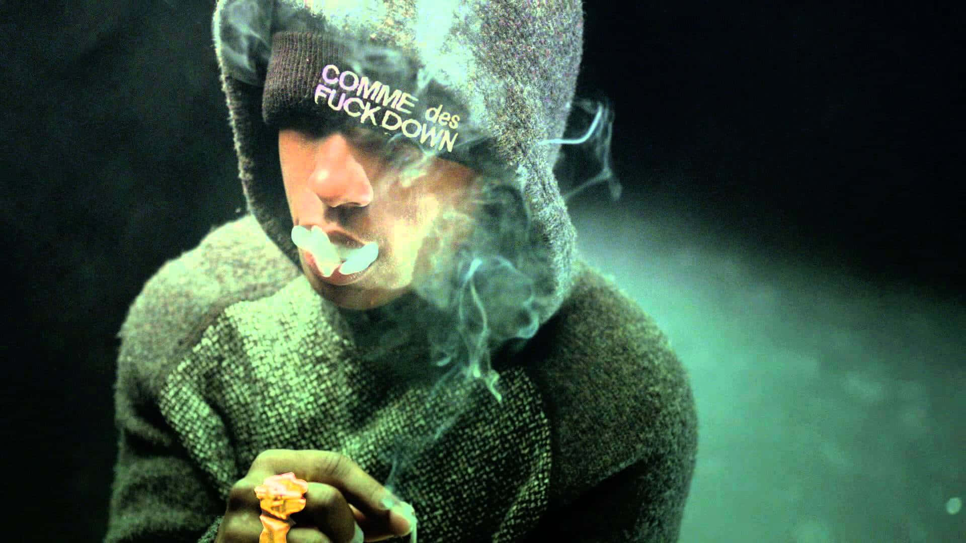 Asap Rocky 吸煙雪茄 壁紙 — 一個穿著連帽衫的人，在深綠色朦朧的背景前，手持點燃的大麻煙並呼出煙霧。