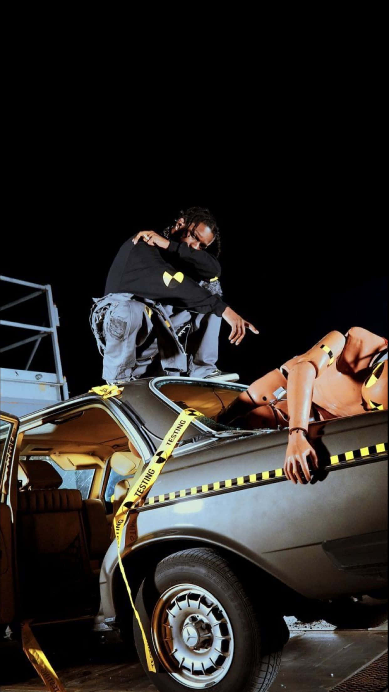 Asap Rocky 登台演出 壁紙 — 一男一女在黑暗夜空下，以戲劇性的姿勢站在一輛被黃色警示膠帶包裹的廢車上。