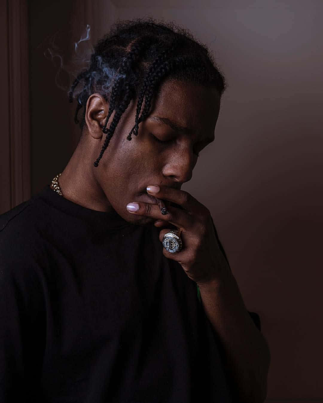 ASAP Rocky 穿著全黑服裝自信站立 壁紙 — 一個辮子男人在昏暗的房間裡拿著香煙呼出煙霧。