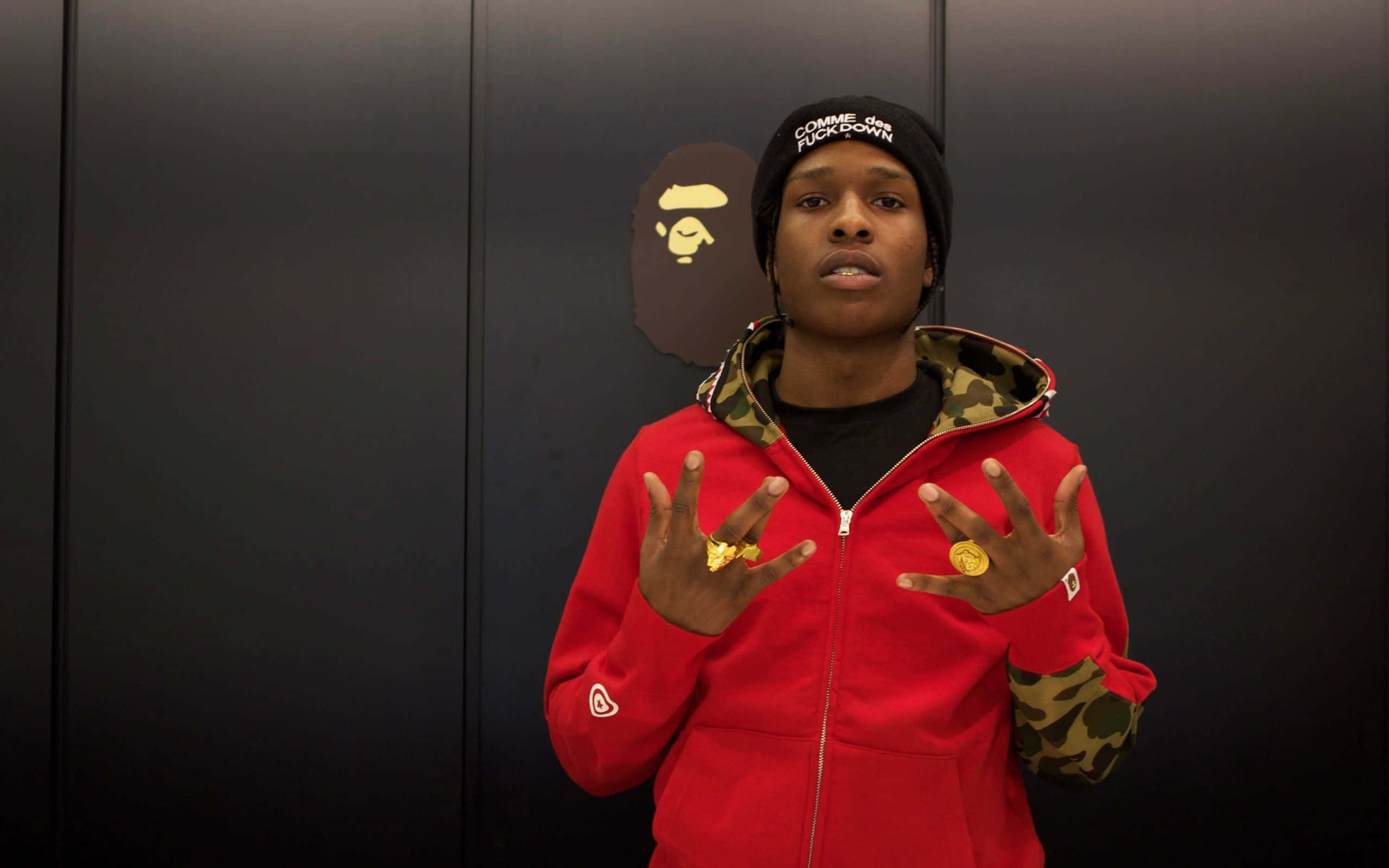 Asap Rocky 走鋼索並演唱 壁紙 — 一個人穿著紅色連帽衫和黑色冷帽，站在帶有BAPE標誌的深色牆壁前，舉起雙手展示金戒指。