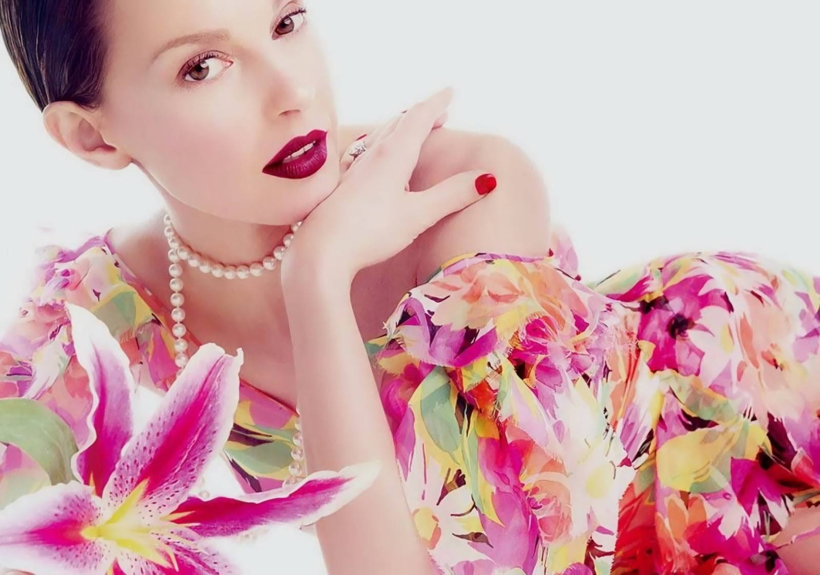 Ashley Judd 紅指甲 壁紙 — 一位擁有鮮豔紅唇和珍珠項鍊的女性，在白色背景前優雅地擺姿勢，身穿鮮豔的花卉連衣裙，前景有一朵大粉紅百合。