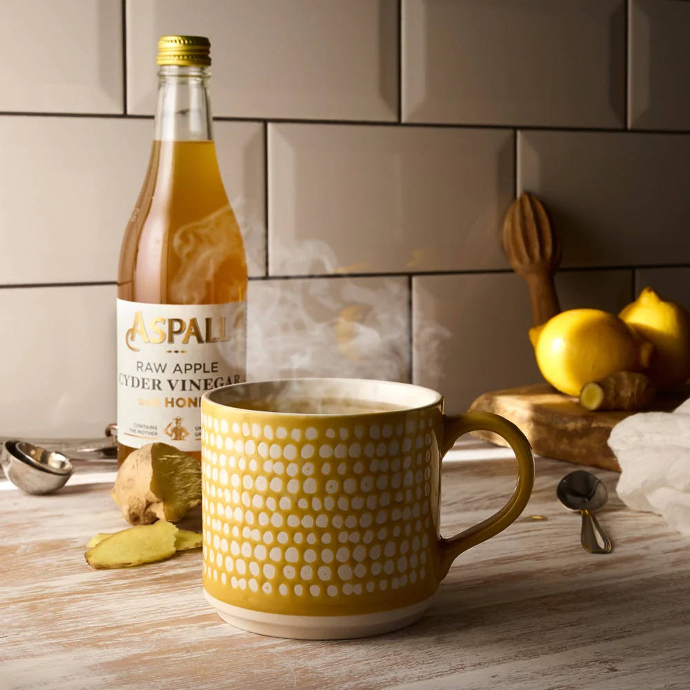 Aspall 純正生蘋果醋 壁紙 — 一個冒著蒸氣的黃色白點馬克杯放在木製枱面上，旁邊有一瓶Aspall蘋果醋檸檬薑，營造舒適的健康飲品。