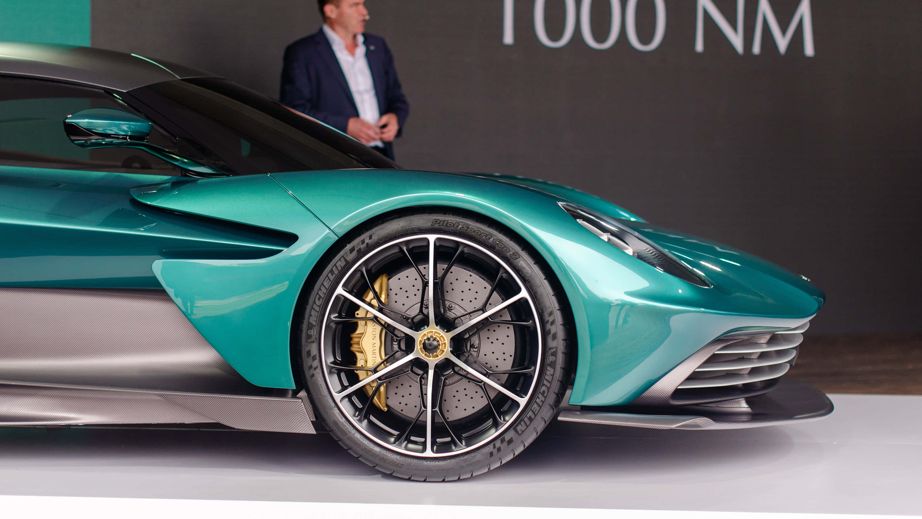 駕馭 Aston Martin，主宰道路 壁紙 — 一輛時尚的藍綠色 Aston Martin 超級跑車配備金色制動卡鉗，在白色平台上於汽車活動中展出。