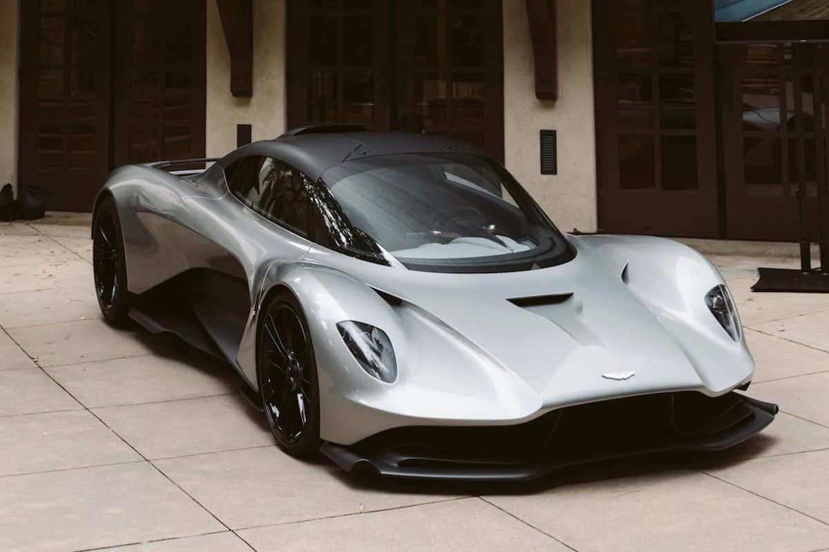 量身訂造 | Aston Martin 壁紙 — 一架時尚的銀色Aston Martin Valkyrie超跑停泊在瓷磚庭院，背景有建築物。
