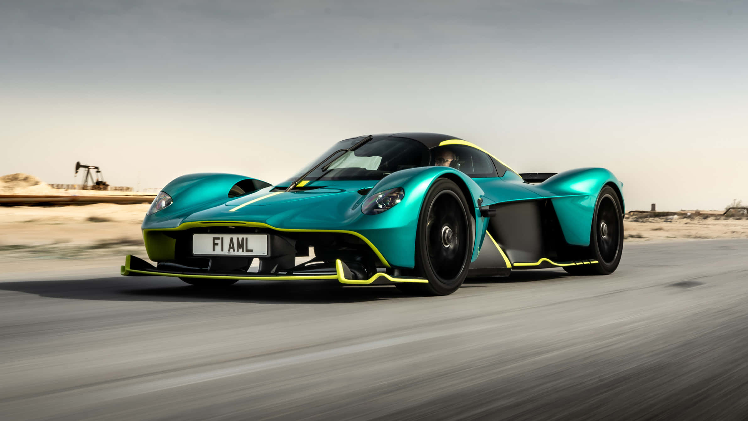 Aston Martin — 無縫奢華 壁紙 — 一輛青綠色和黃色的Aston Martin Valkyrie hypercar在沙漠公路上飛馳，背景有石油鑽井平台。