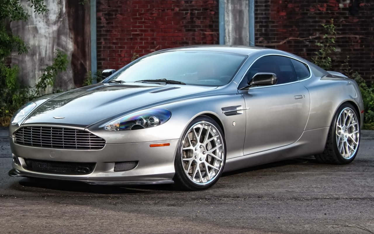 Aston Martin DB9 - 力量與設計的完美融合 壁紙 — 一架時尚的銀色Aston Martin跑車停泊在瀝青路上，旁邊是帶有綠色植物的風化磚牆。