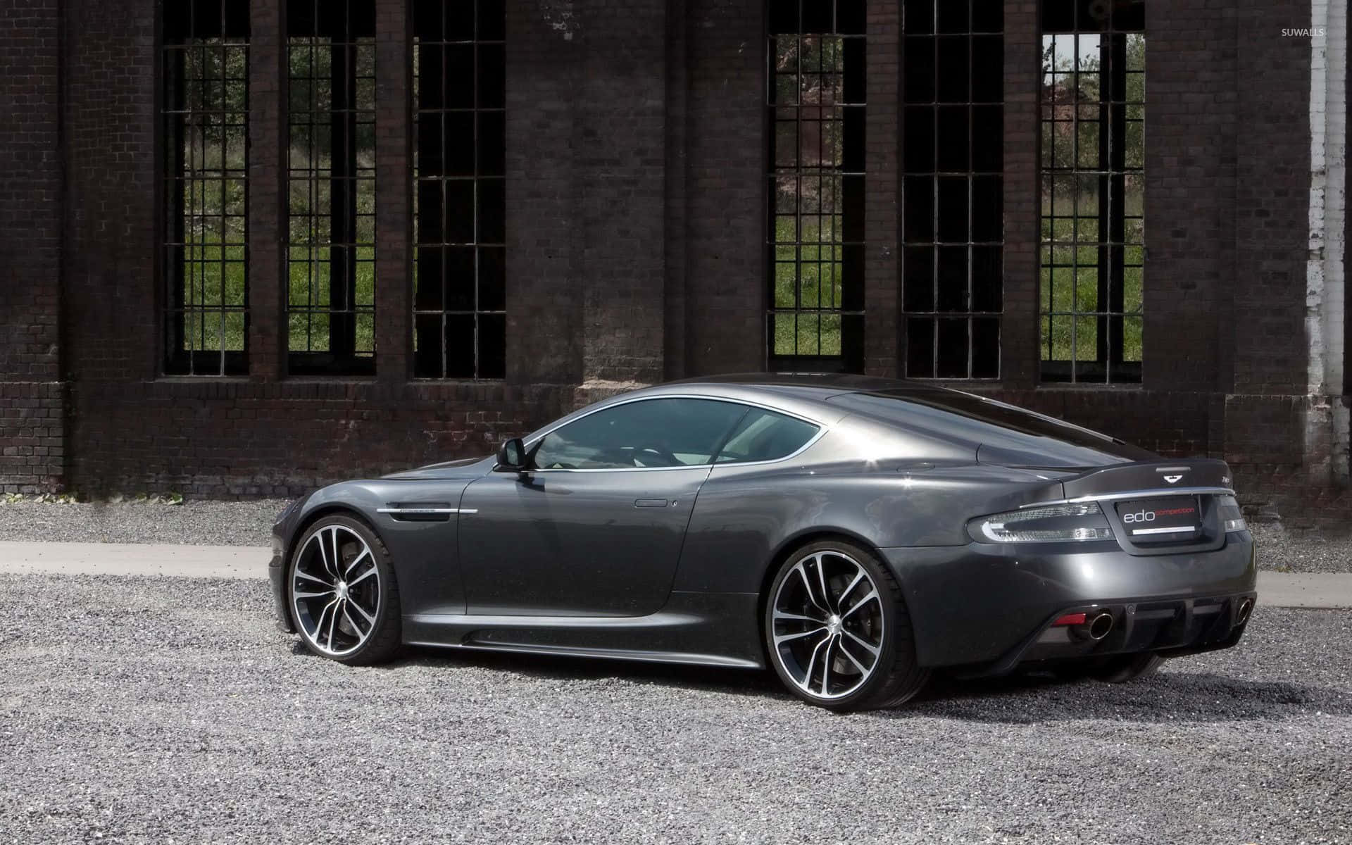 Aston Martin DB9 優雅與力量 壁紙 — 一架光滑的啞光灰色 Aston Martin 跑車停泊在碎石路上，前面是一棟帶有高窗的風化磚砌建築。
