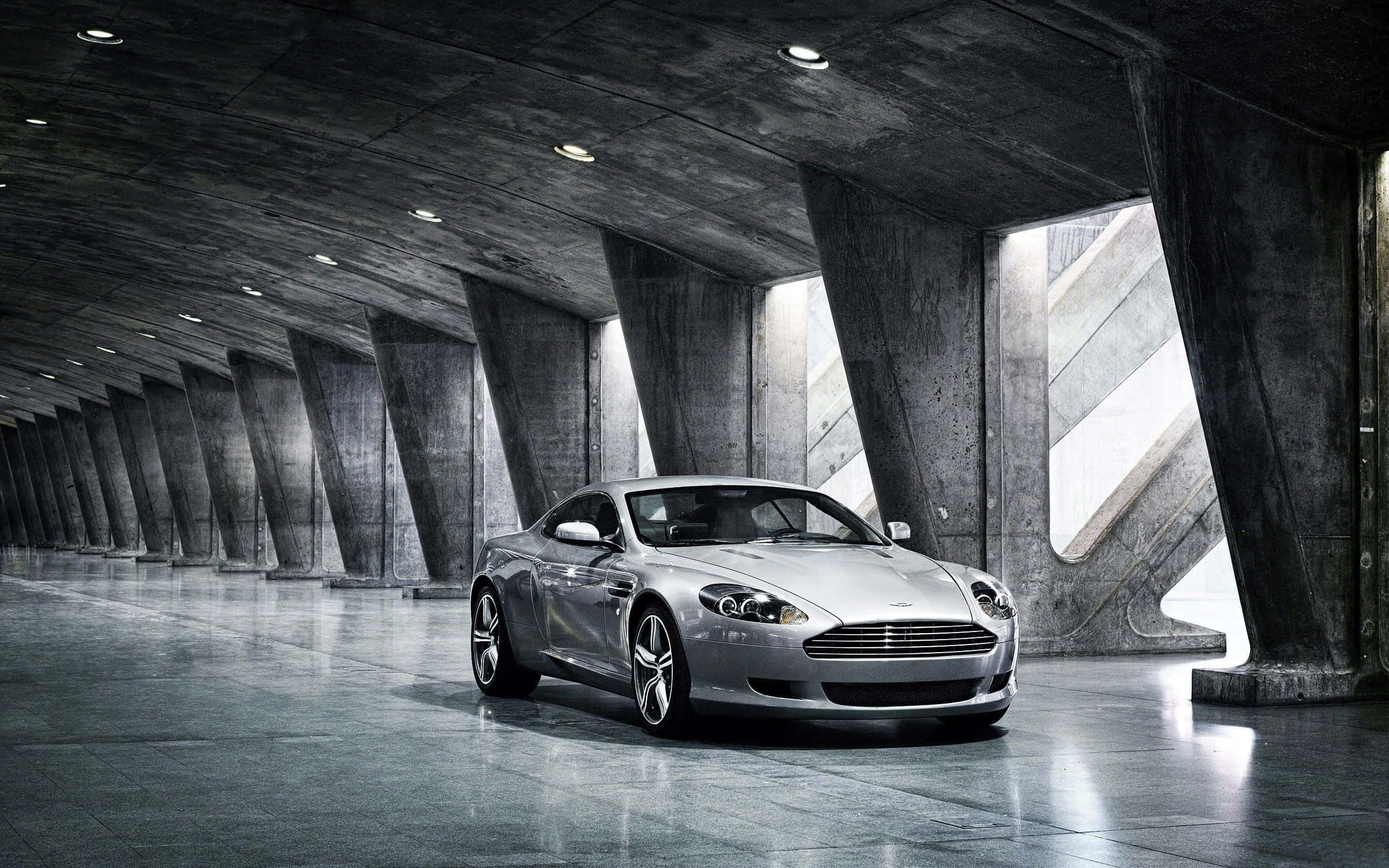 Aston Martin DB9穿梭風景城市景觀 壁紙 — 一輛時尚的銀色跑車在寬敞的混凝土拱形車庫中，在工業燈光下閃閃發光。