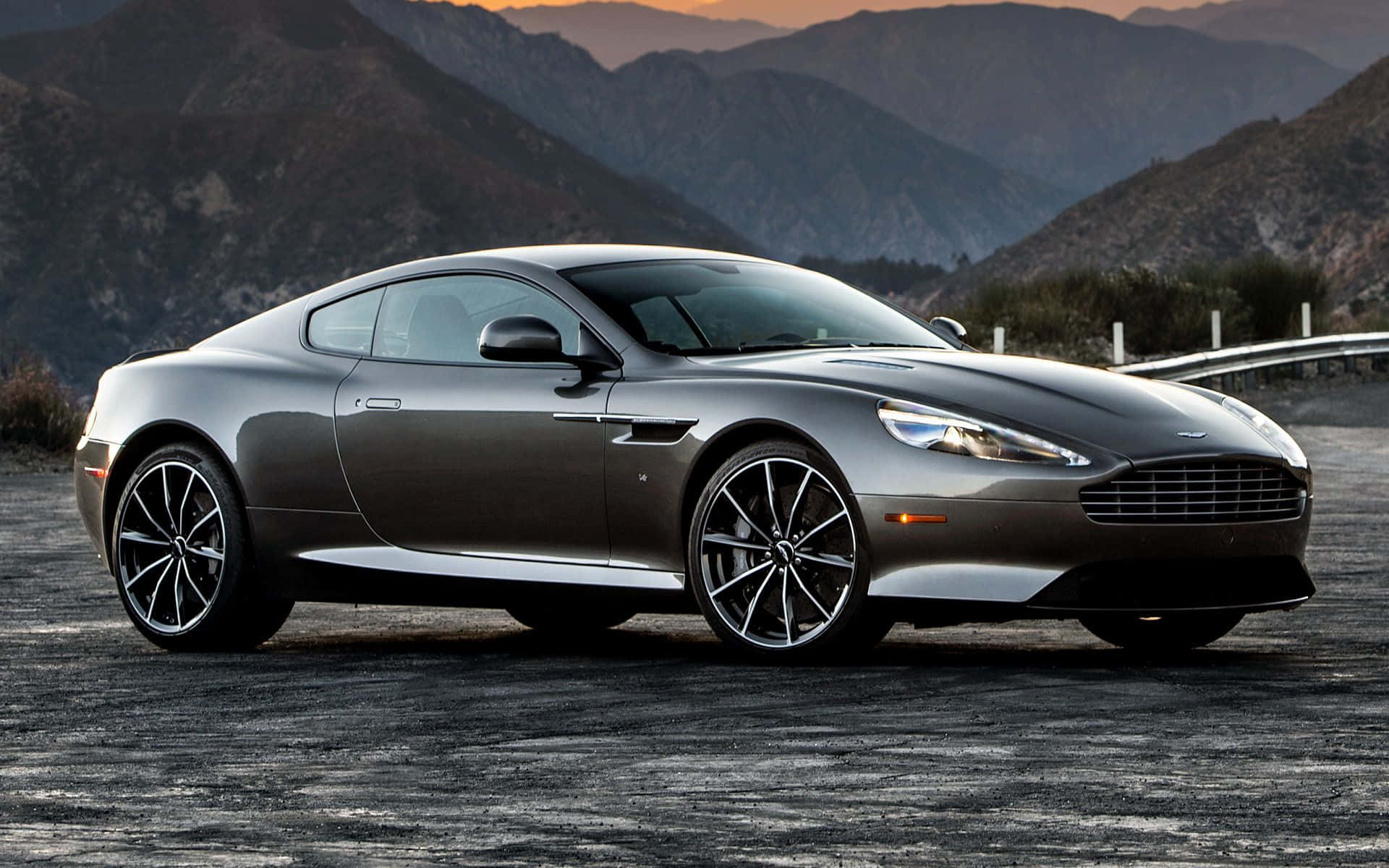 驚艷的Aston Martin DB9於優雅場景中 壁紙 — 一架流線型銀色Aston Martin跑車停泊在日落時分的山路上，背景是崎嶇的山峰。