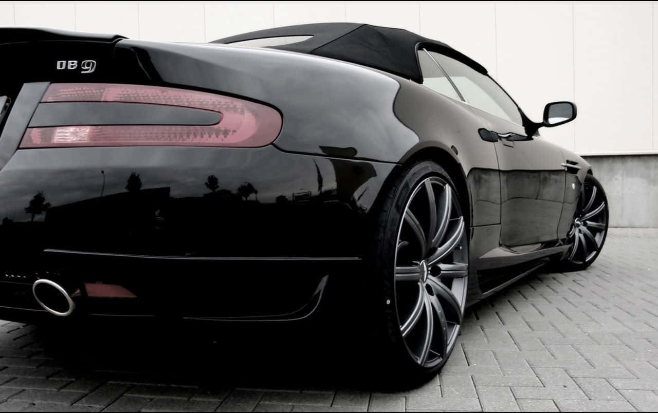 銀色 Aston Martin DB9 馳騁公路 壁紙 — 一架光滑的黑色Aston Martin DB9開篷跑車停泊在鋪砌路面上，其光澤表面反射著周圍環境。