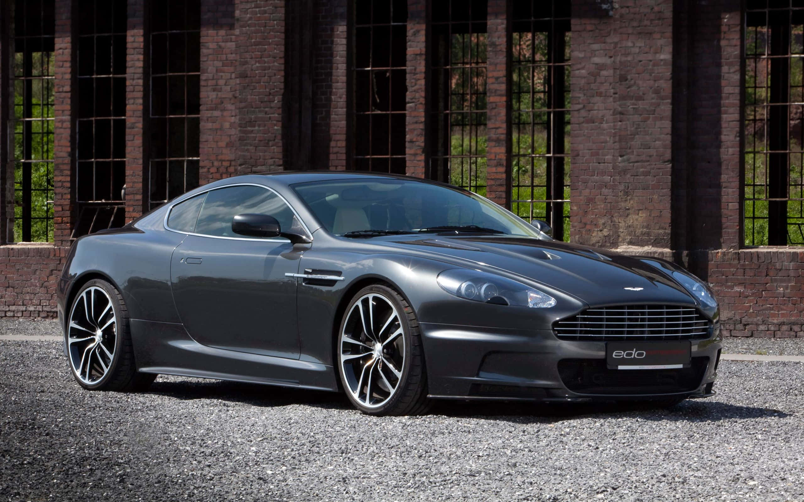 賽道上令人驚嘆嘅銀色Aston Martin DB9 壁紙 — 一架型格啞灰色Aston Martin跑車停泊在碎石路上，前方是帶有高窗的風化磚建築。